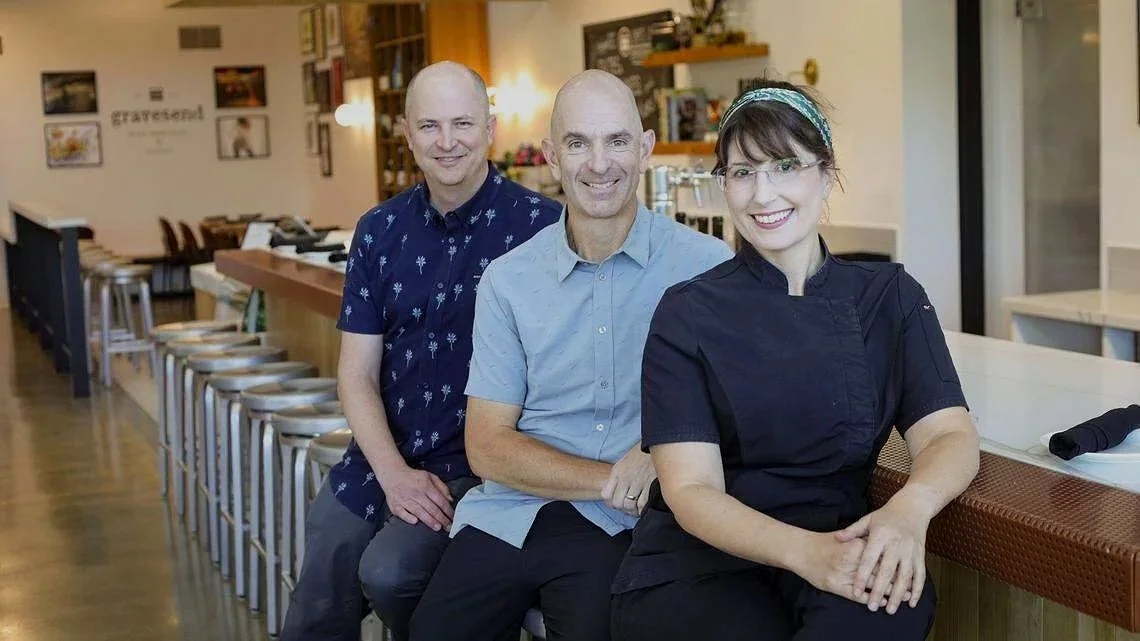 Three people sitting side by side in a restaurant or cafe, smiling at the camera.