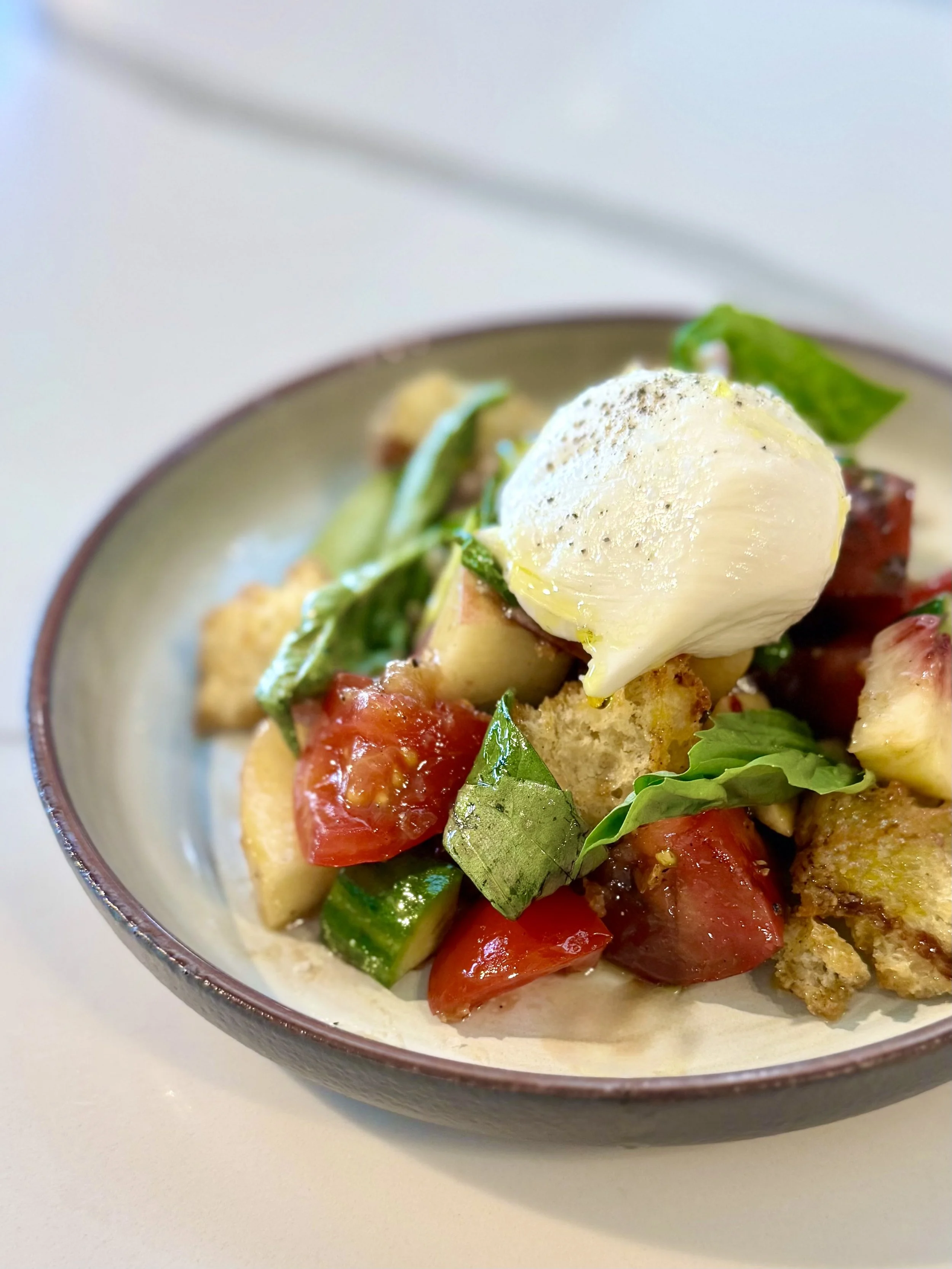 A close-up of a salad dish in a bowl containing cherry tomatoes, green lettuce, slices of avocado, and a poached egg with pepper on top.