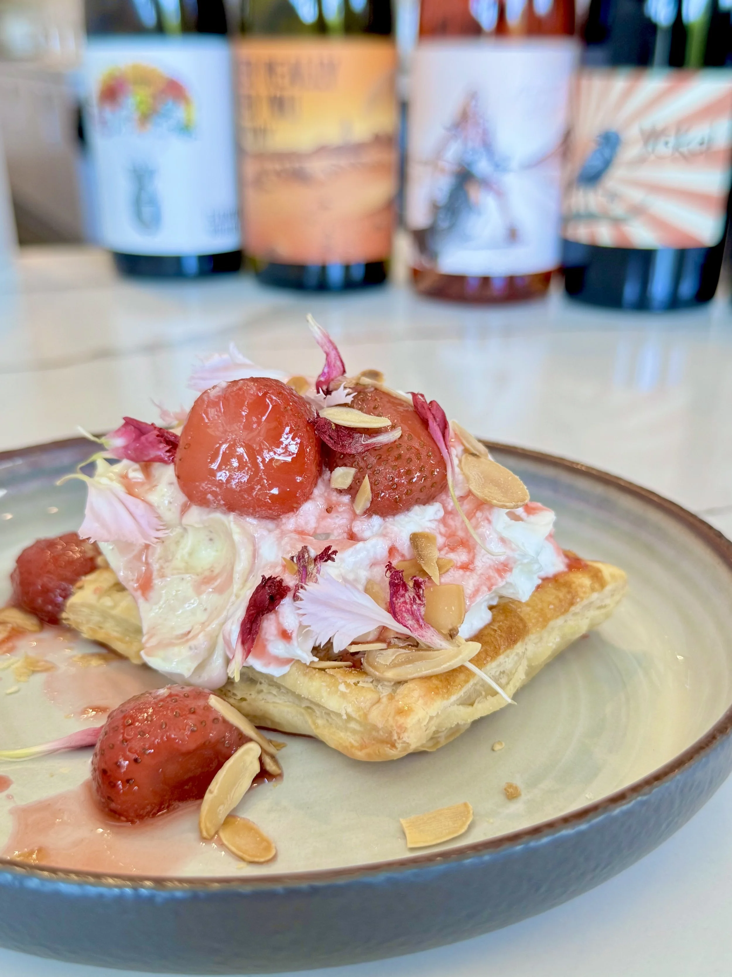 A close-up of a piece of strawberry tart topped with sliced strawberries, slivered almonds, and flower petals in a blue ceramic bowl.