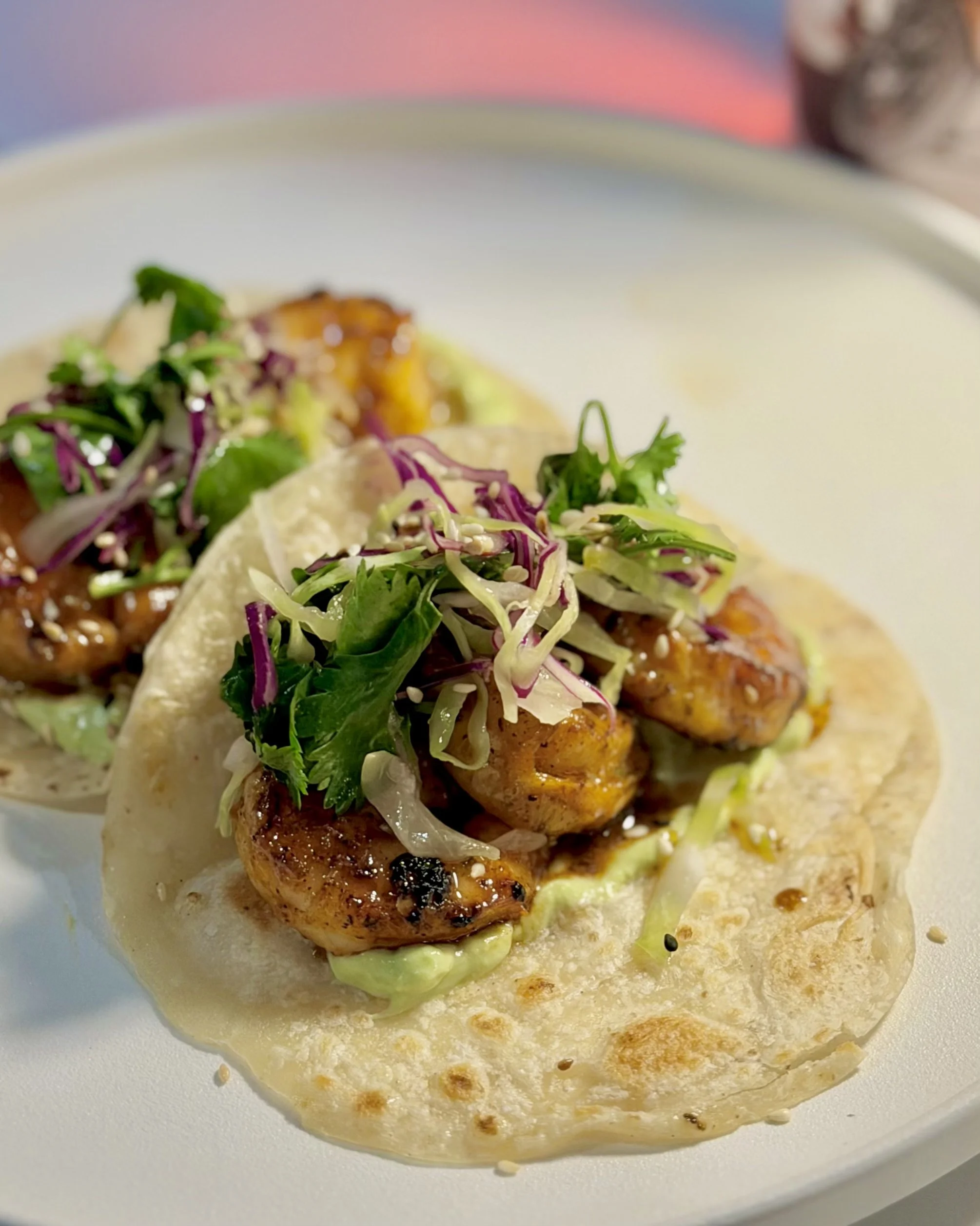 Close-up of two Asian-style tacos on a white plate, filled with grilled chicken, shredded cabbage, cilantro, and sesame seeds.