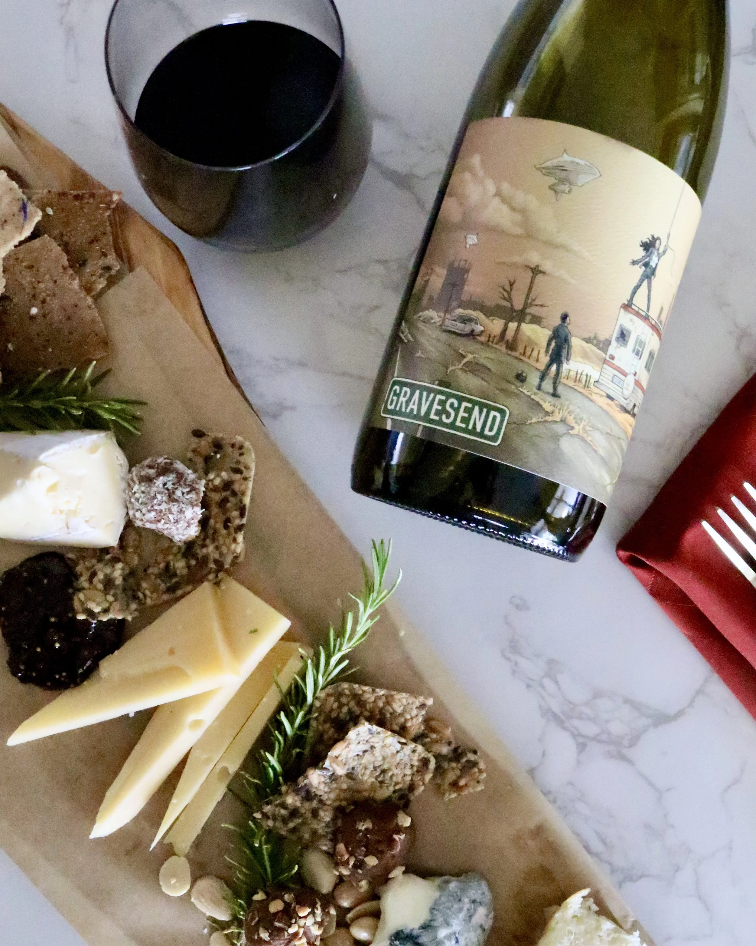 A cheese and charcuterie board with various cheeses, nuts, and herbs, alongside a glass of red wine and a bottle with artistic label painting a landscape scene.
