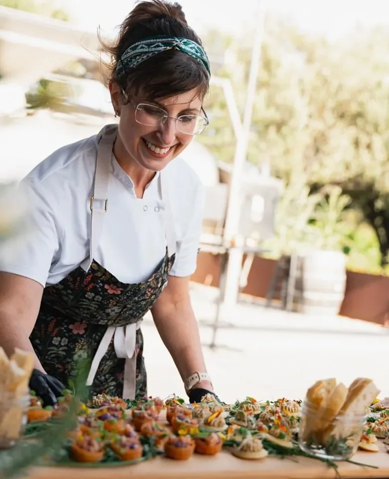 A woman wearing glasses, a headscarf, and a floral apron smiles as she arranges colorful appetizers and mini sandwiches on a table outdoors.