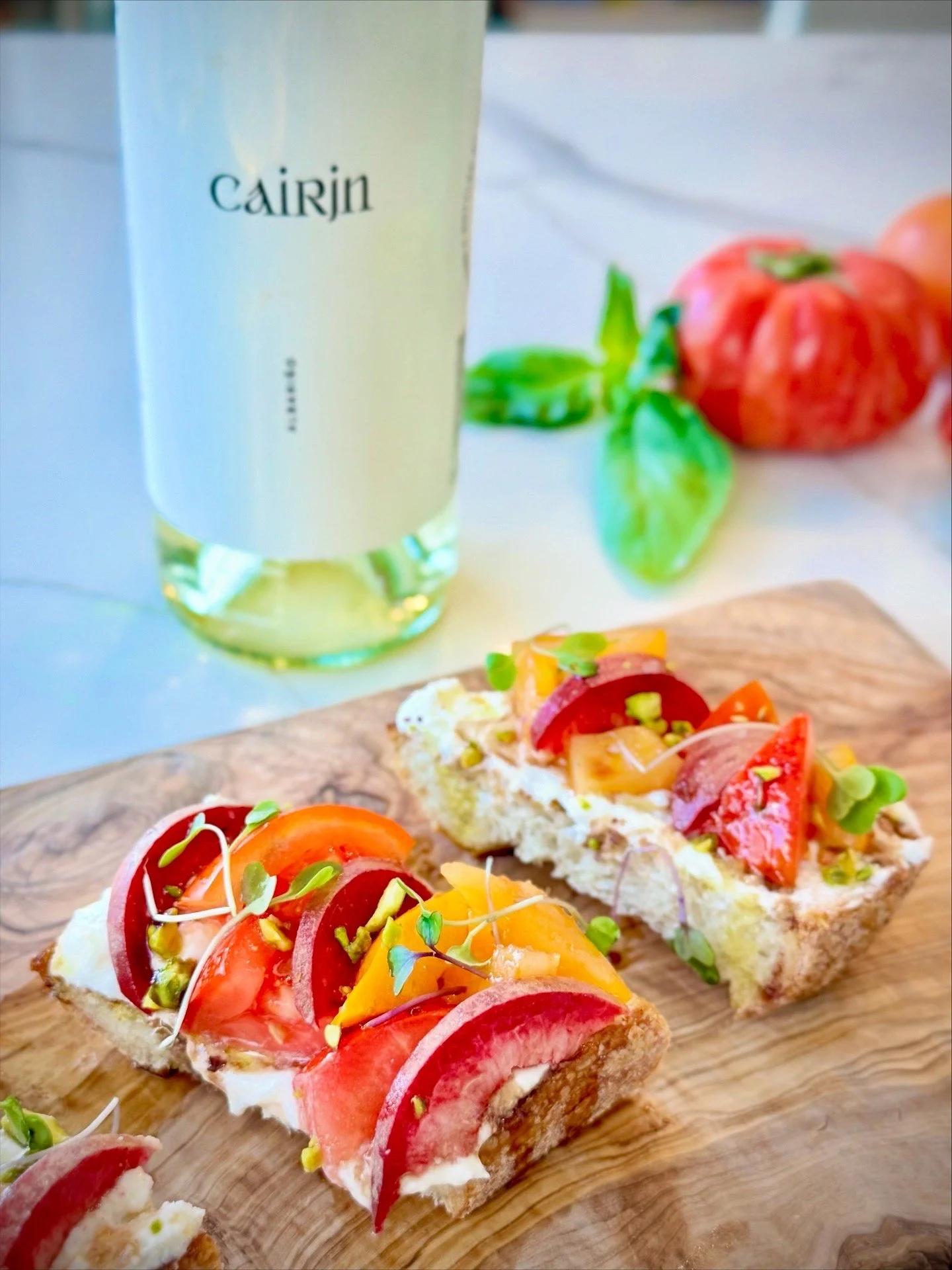 Open-faced sandwiches with tomato slices, microgreens, and cheese on crusty bread, served on a wooden board, with a bottle of Cairin beverage and some tomatoes in the background.