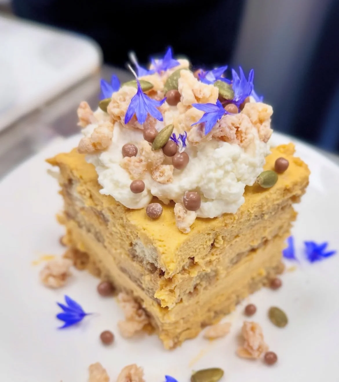 A square piece of layered dessert topped with whipped cream, edible blue flowers, crunchy crumble, green pumpkin seeds, and small round chocolate and frosting sprinkles.