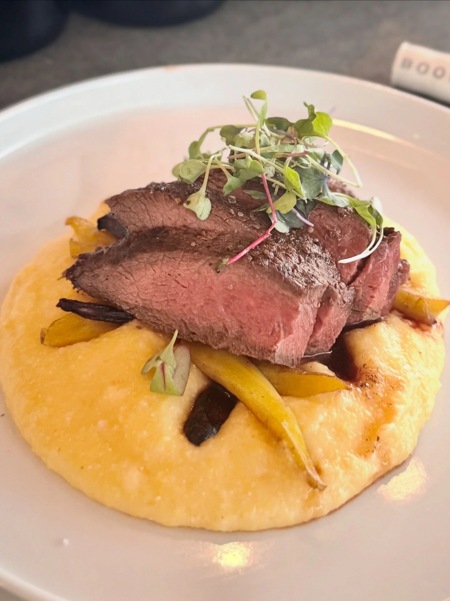 Close-up of a plate with mashed potatoes topped with sliced roasted beef, grilled vegetables, and microgreens.