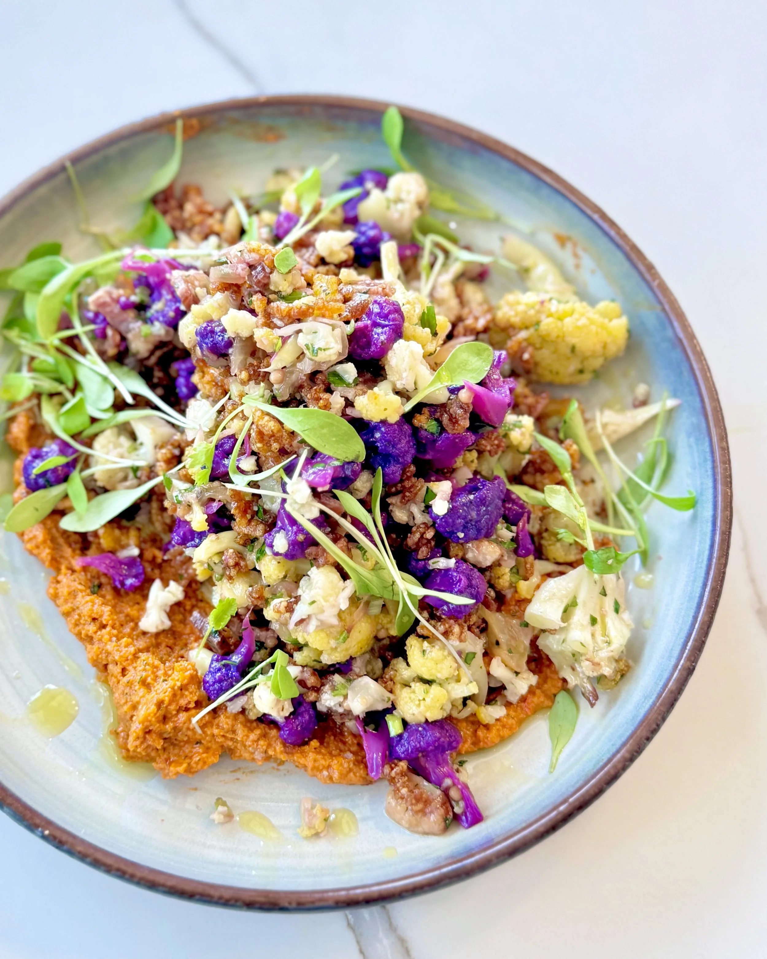 A bowl of food featuring a breaded fish fillet topped with purple and green microgreens and a mix of crumbled cheese, purple cabbage, and other vegetables.