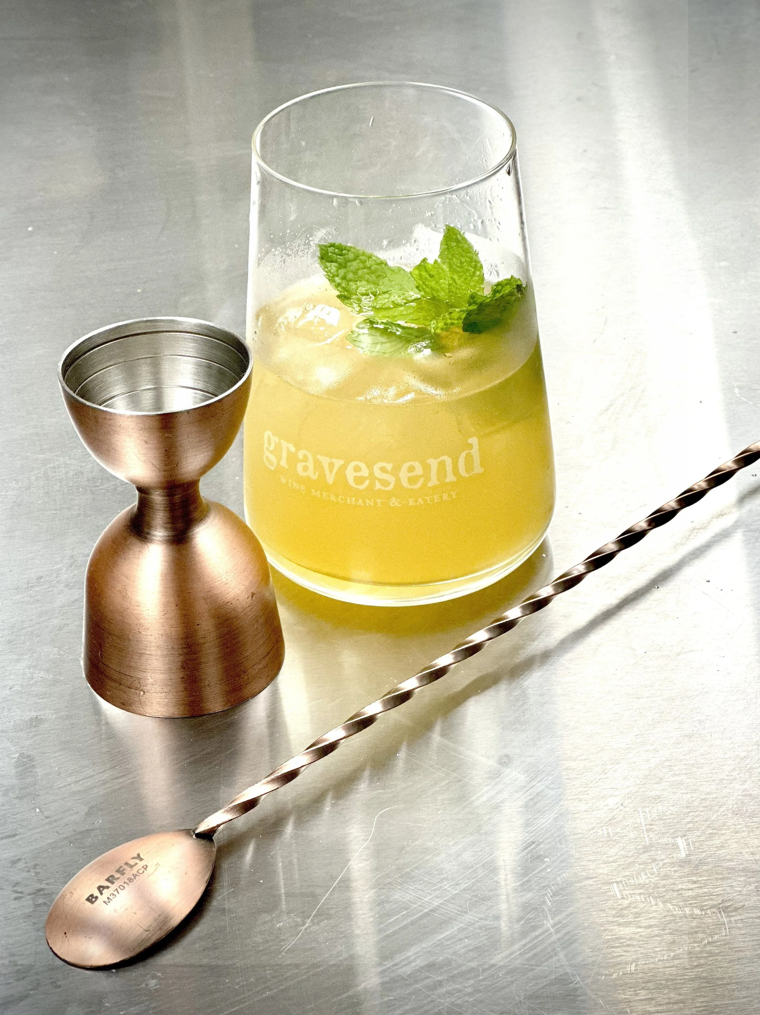 A metallic spoon on a stainless steel surface, with a glass of cold beverage garnished with mint leaves and ice. The glass has text that reads "gravesend WINE MERCHANT & EATERY."