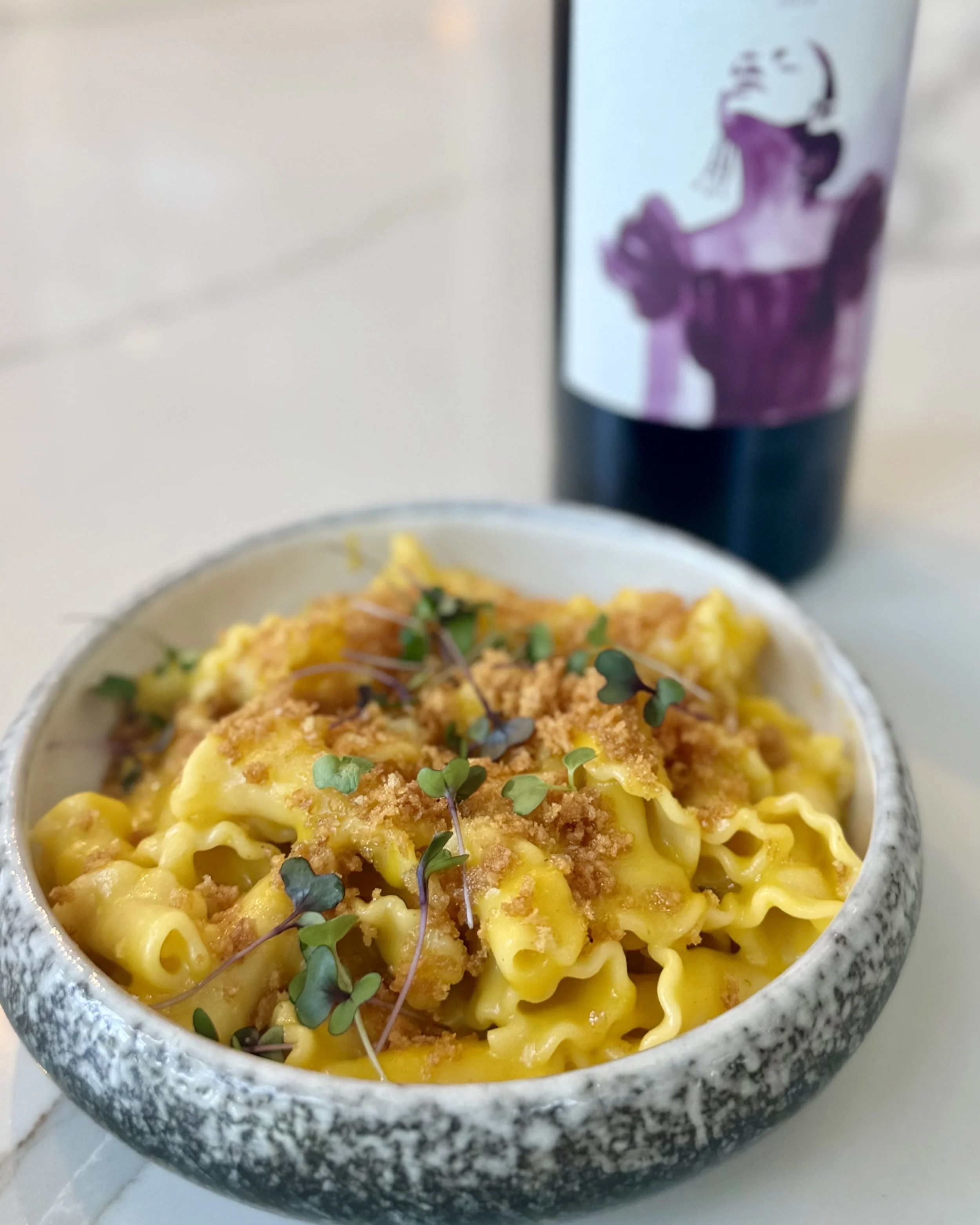 A bowl of cheesy pasta topped with bread crumbs and microgreens, with a purple and white soda cup in the background.