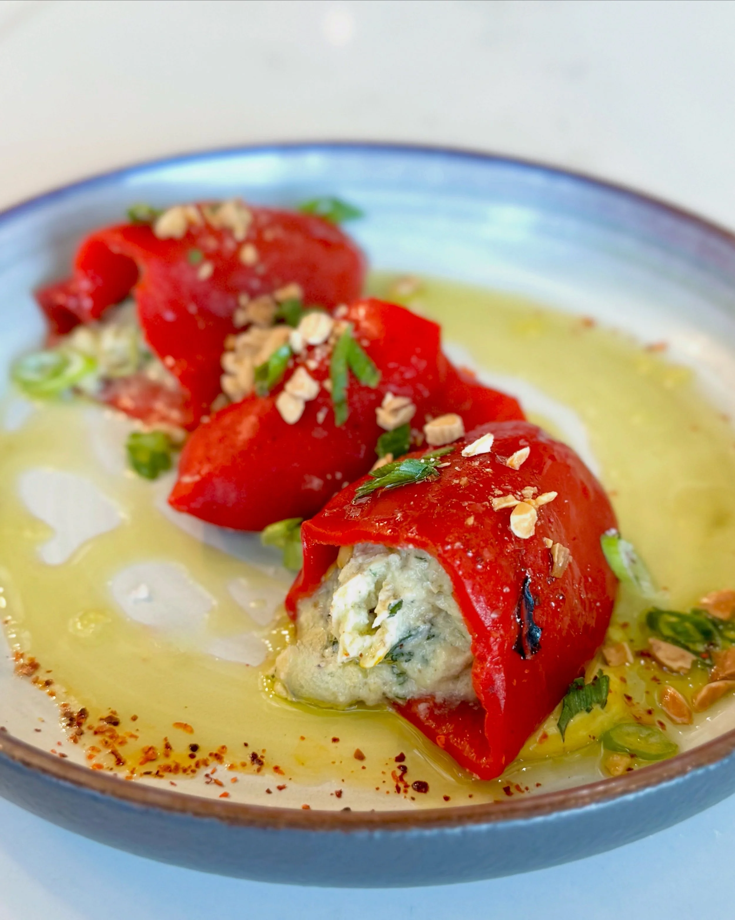 Close-up of a dish with stuffed red peppers, garnished with chopped green onions and chopped nuts, in a yellow sauce with red pepper flakes on the edge of the plate.