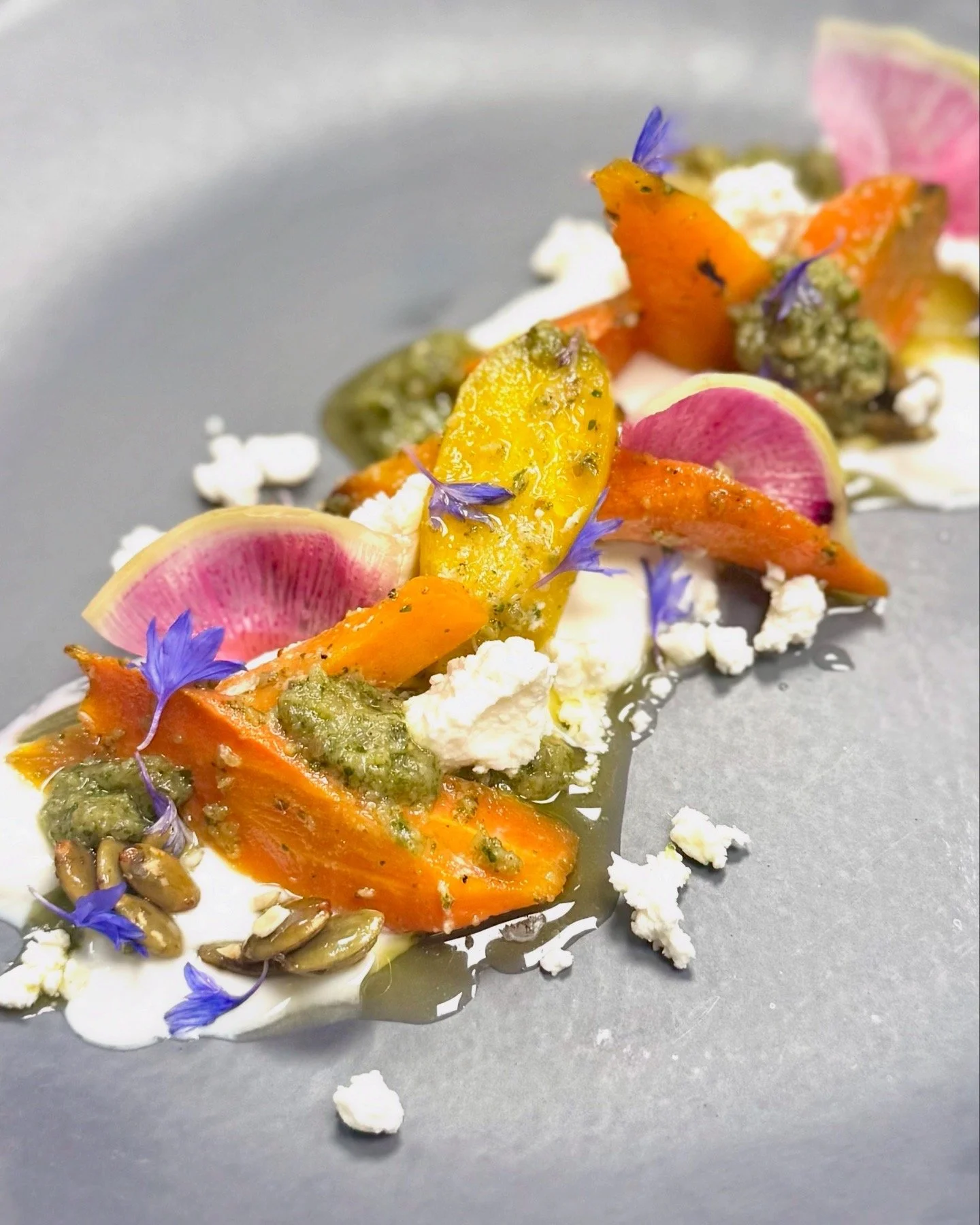 Close-up of a gourmet dish with roasted orange vegetables, pink radish slices, purple edible flowers, green pesto, crumbled white cheese, sunflower seeds, and a drizzle of sauce on a gray background.
