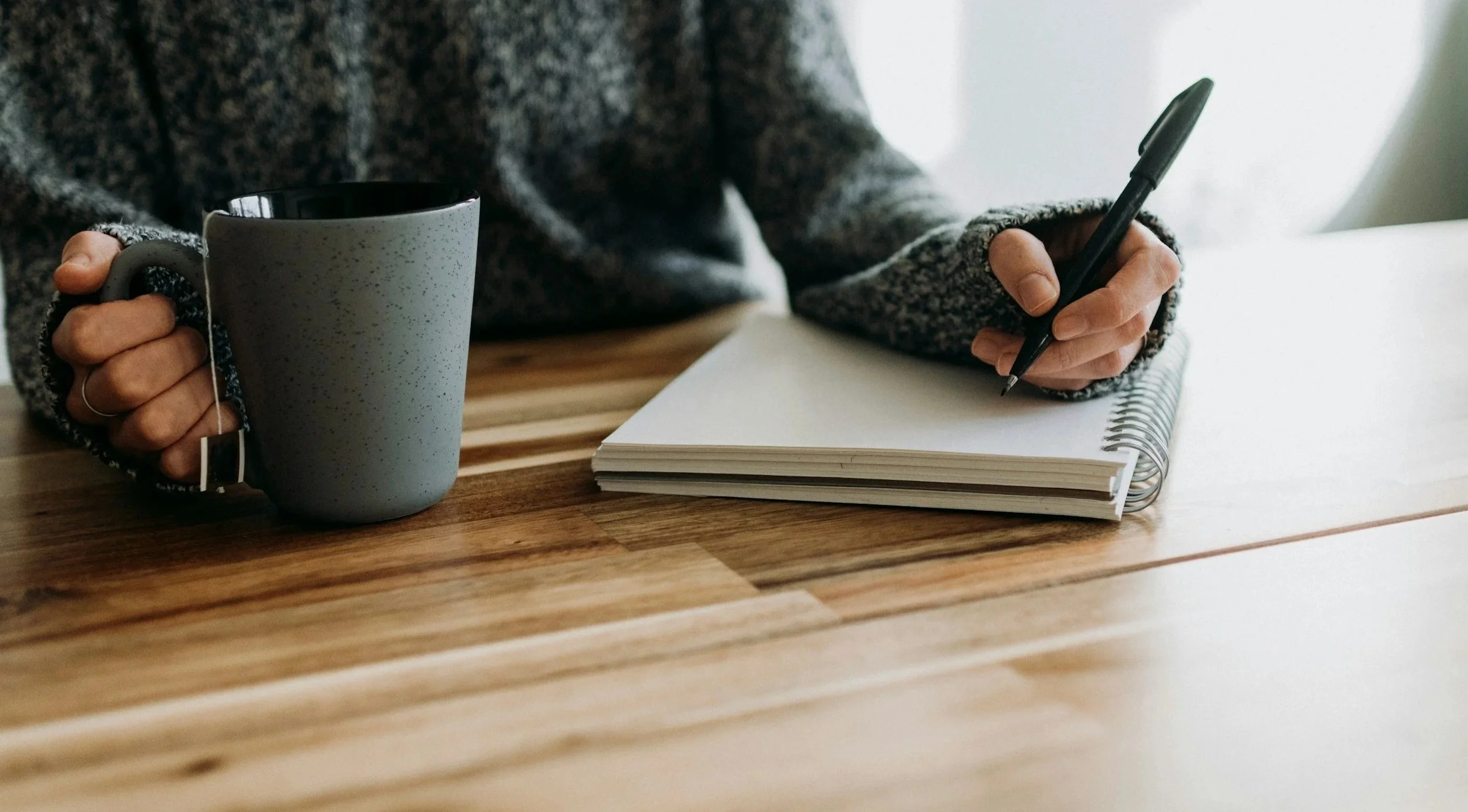 Person with a fuzzy sweater holding a gray mug in one hand and writing in a spiral notebook with a black pen on a wooden table.