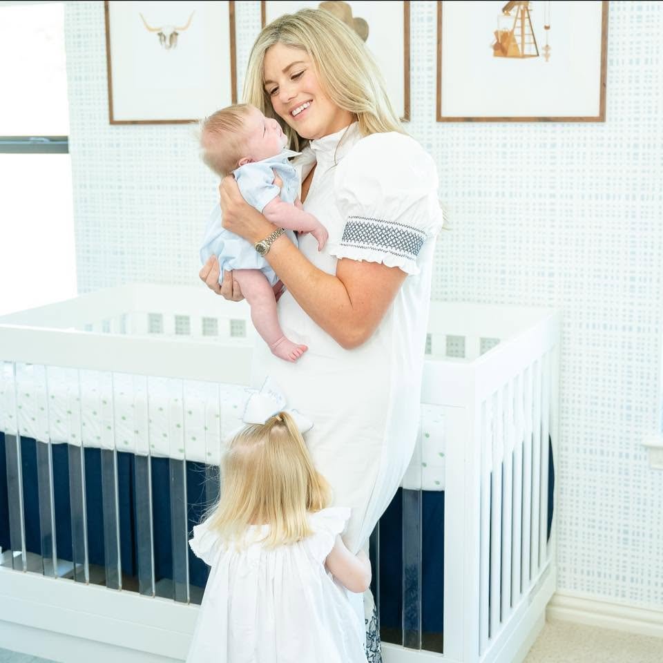 A woman, possibly a mother, holding a sleeping baby in a nursery. A young girl with blonde hair, wearing a white dress, is standing close to her.