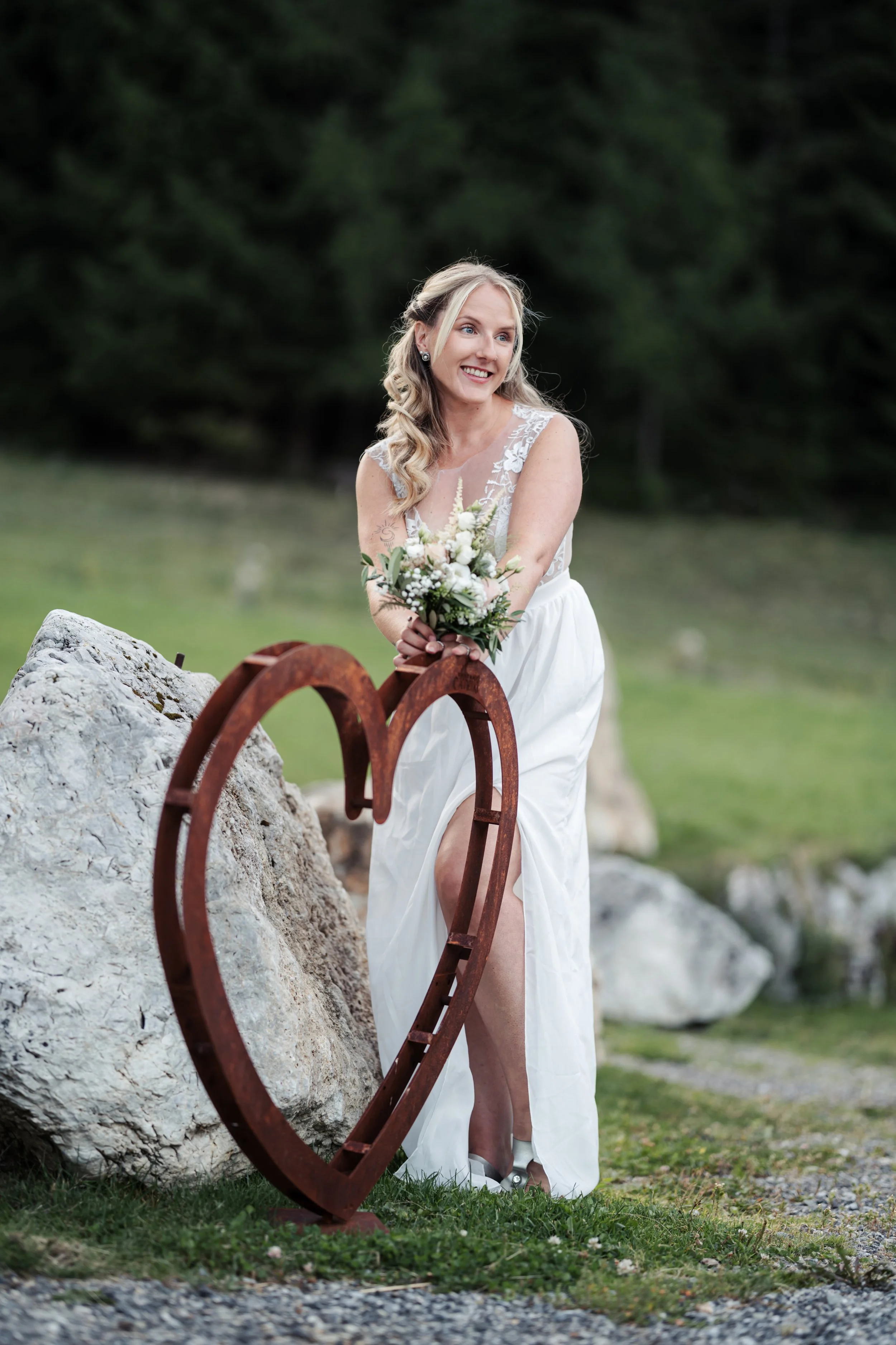 Eine lächelnde Frau in einem weißen Kleid, die im Freien auf einem Pfad steht, hält einen Blumenstrauß, während sie einen rustikalen Metallherz-Schmiedeeisenskulptur berührt.
