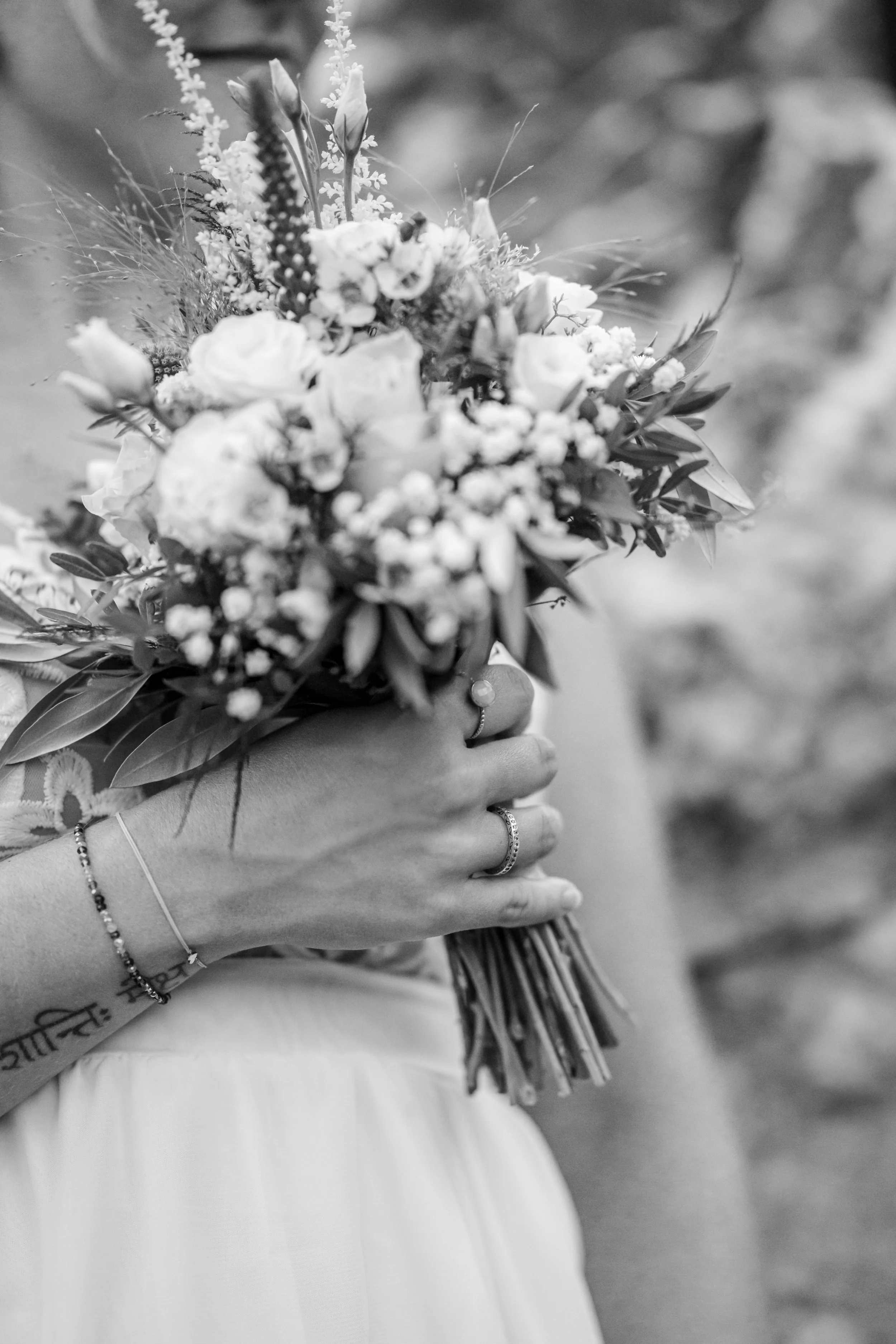 Hand hält einen Brautstrauß mit Blumen und Gräsern. Person trägt Schmuck an Hand und Arm, sichtbare Tätowierung am Handgelenk, Kleidung in Weiß. Hintergrund unscharf.