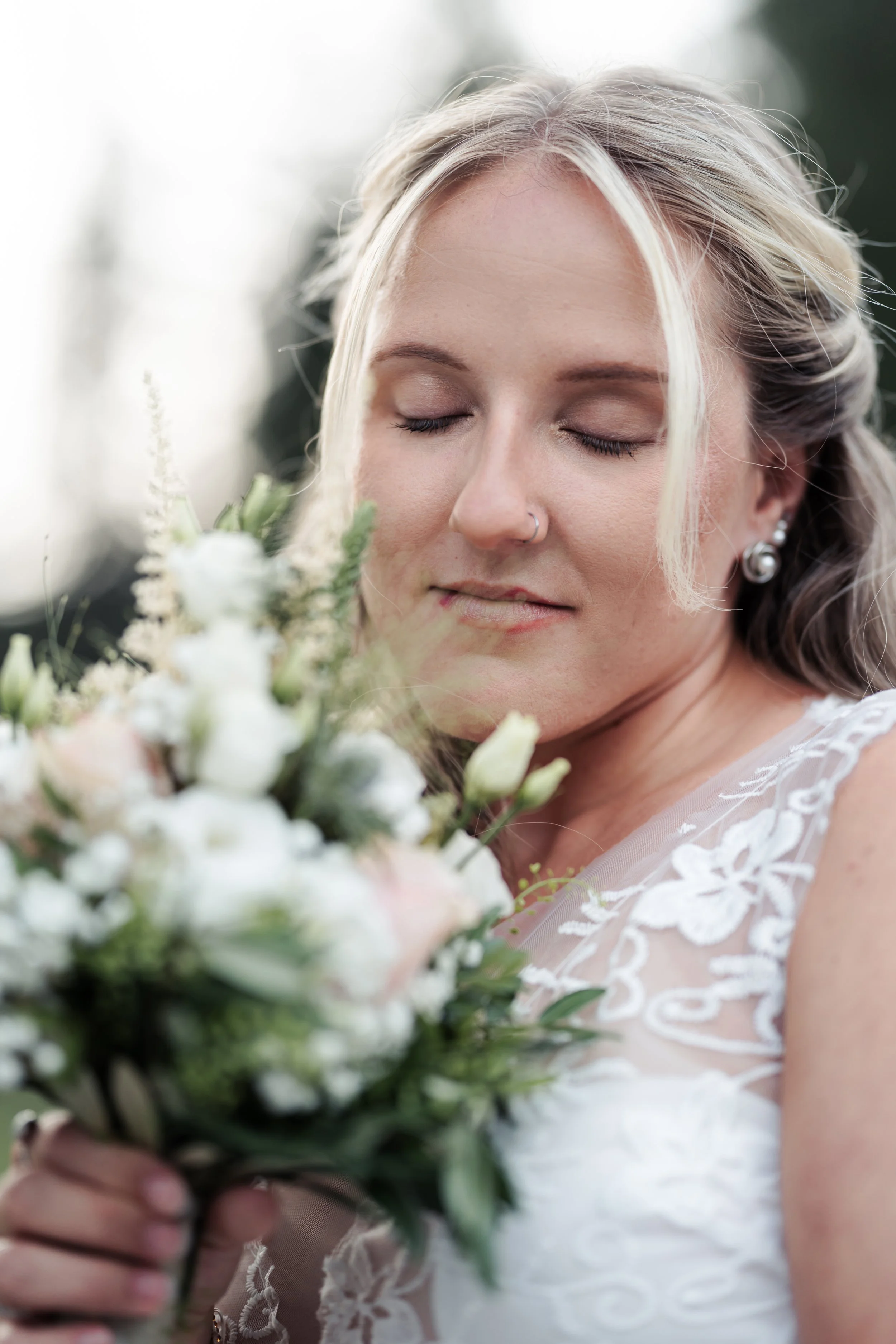 Eine glückliche Frau mit blonden, gewellten Haaren, die eine weiße Spitzenbrautkleid trägt, hält einen Blumenstrauß mit weißen und grünen Blumen. Sie hat geschlossene Augen und einen Nasenringe. Die Szene wirkt romantisch und feierlich.