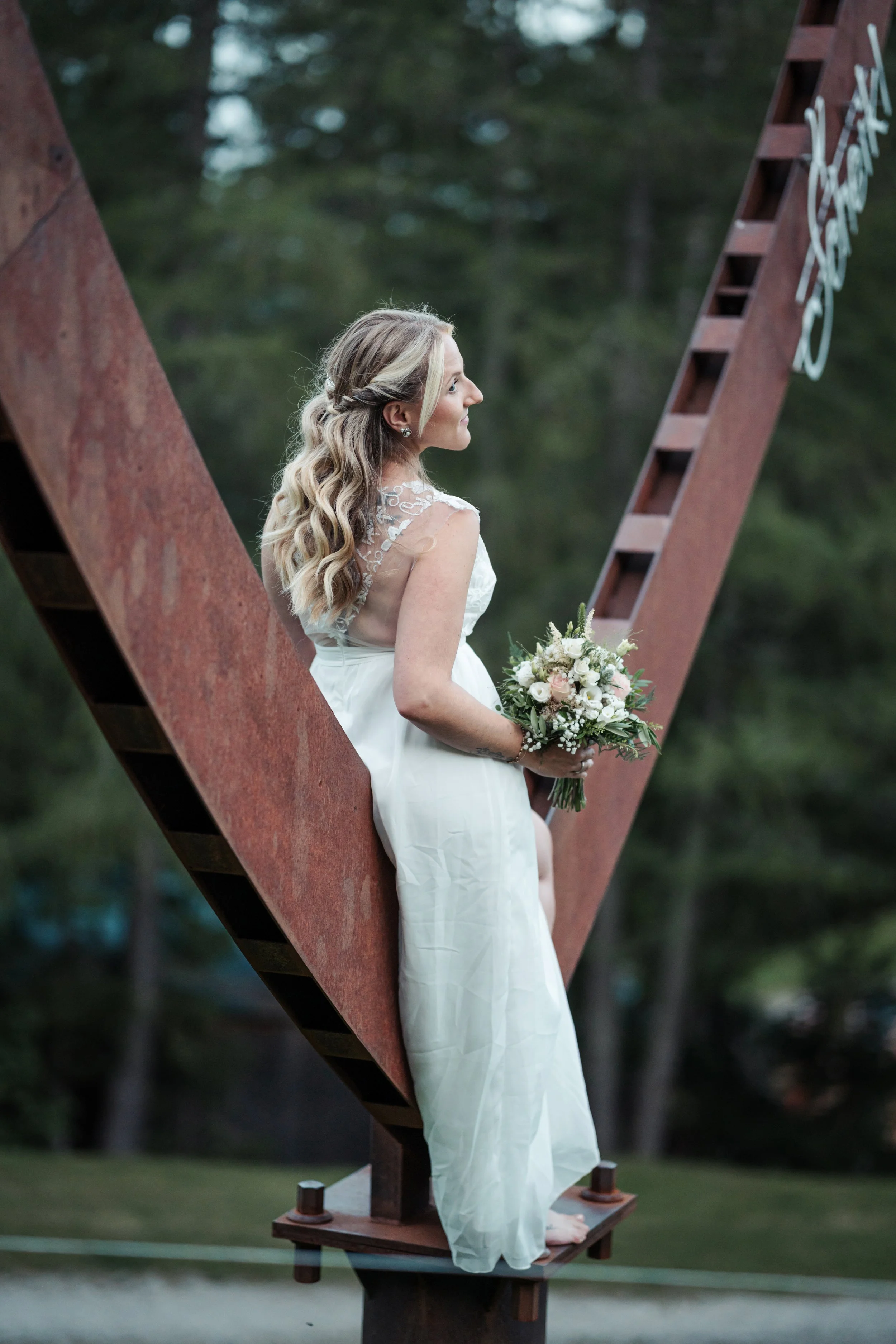 Eine Frau in einem weißen Brautkleid hält einen Blumenstrauß und steht an einer großen Metallskulptur im Freien mit Bäumen im Hintergrund.