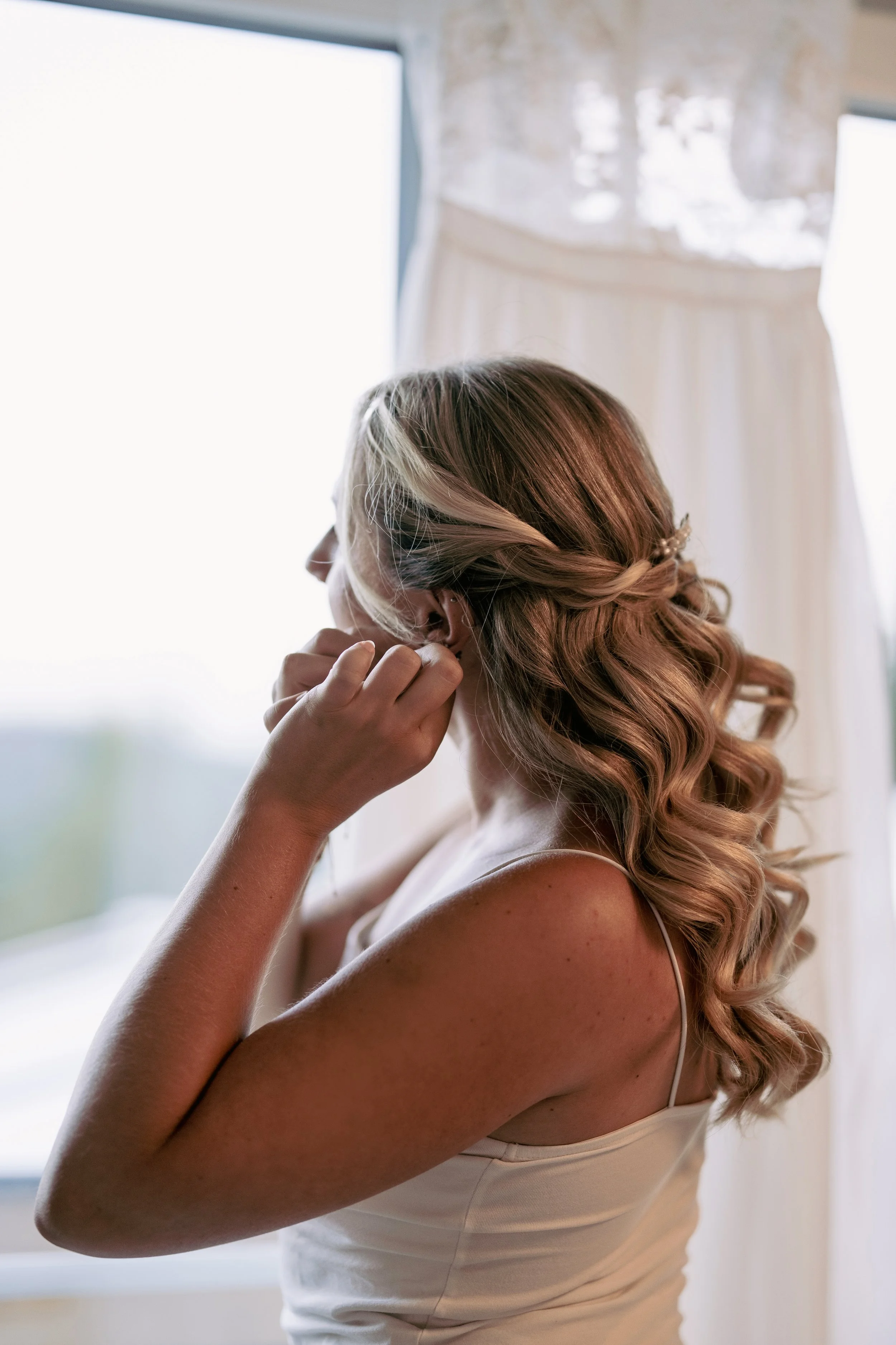 Eine Frau mit lockigem, hellbraunem Haar trägt einen weißen, trägerlosen Dress und richtet sich die Ohrringe. Das natürliche Licht kommt durch ein großes Fenster im Hintergrund.