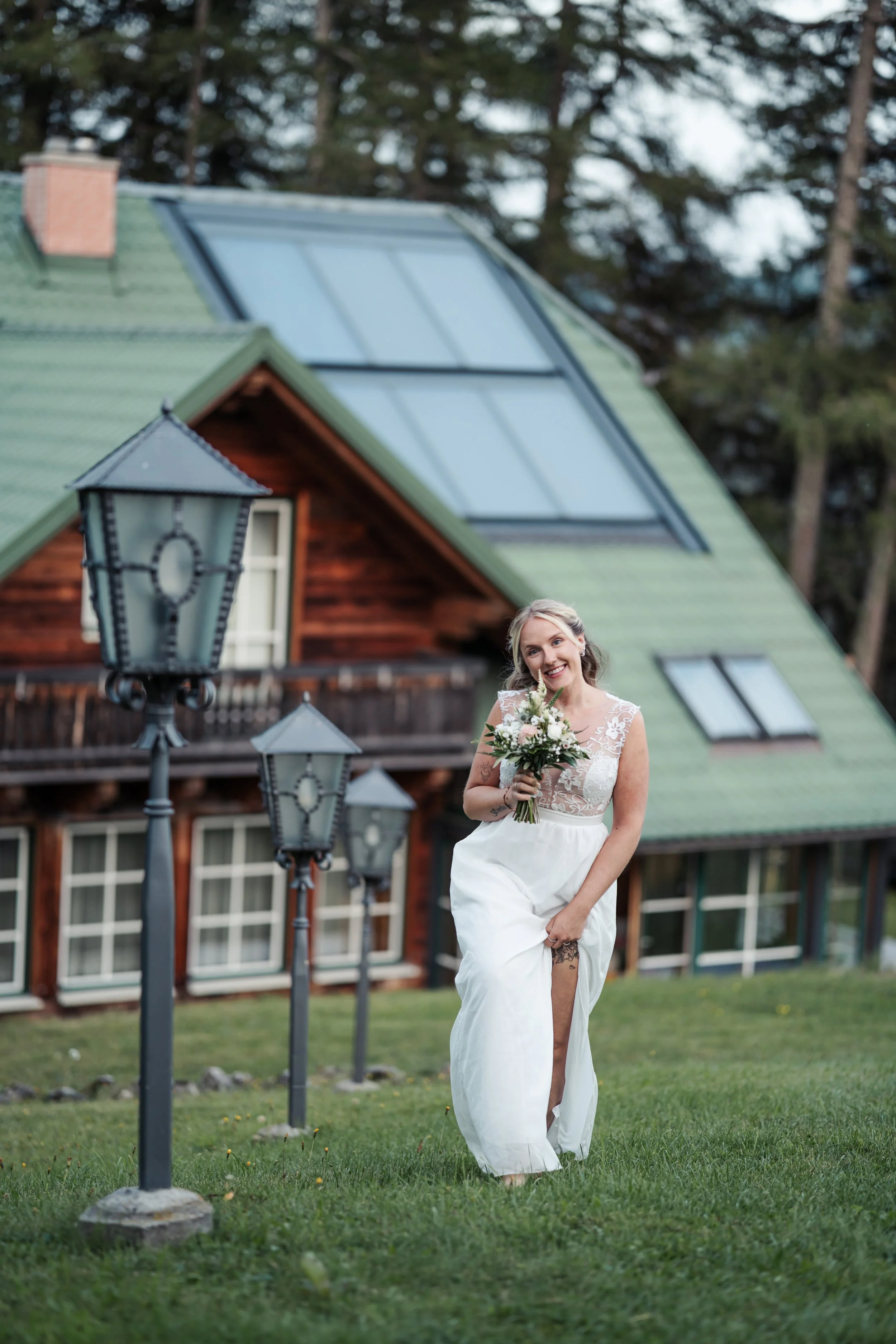 Eine lachende Frau in einem weißen Brautkleid mit Blumenstrauß im Freien vor einem Holzhaus mit grüner Dachdeckung. Im Vordergrund sind mehrere alte Laternen zu sehen, die entlang eines Weges aufgestellt sind, umgeben von grünem Rasen.