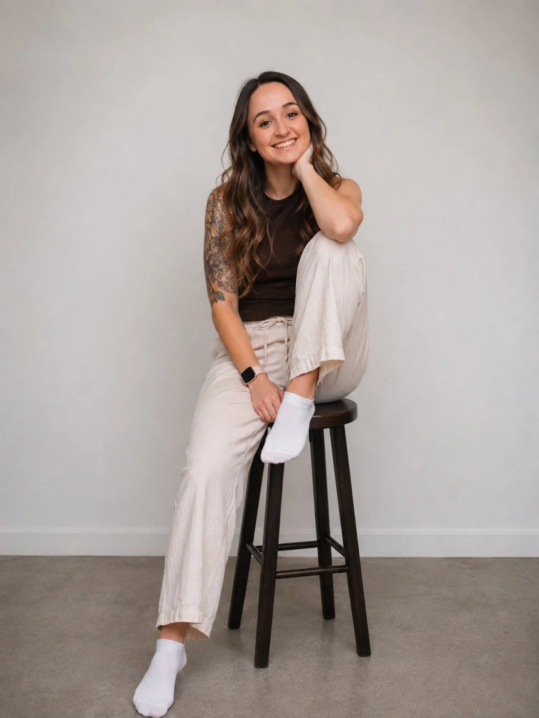 Junge Frau mit Tattoo auf dem linken Arm sitzt auf einem Barhocker vor einer weißen Wand, trägt eine schwarze Top, helle Hosen und weiße Socken, lächelt, hat lange braune Haare.