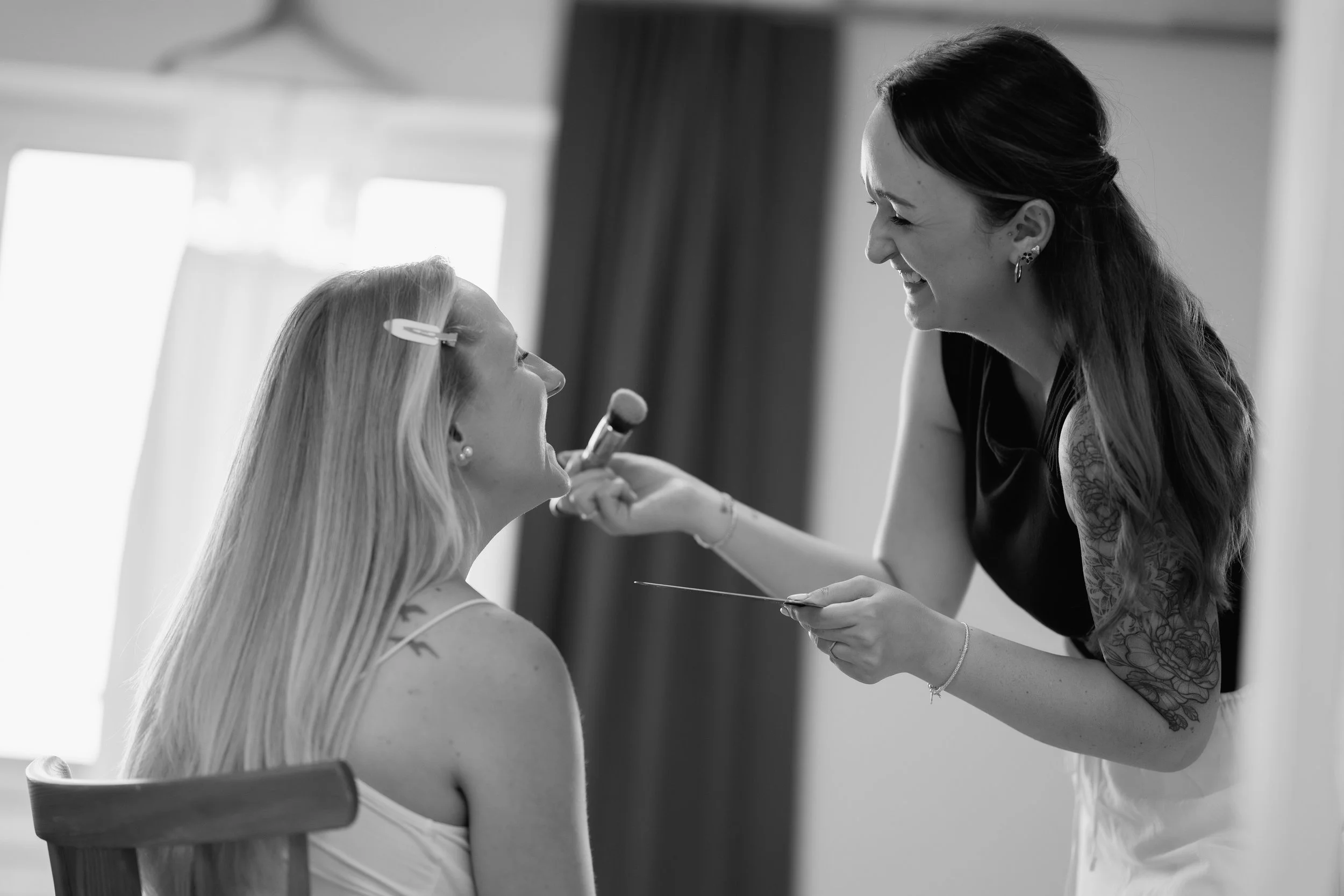 Eine Frau beim Make-up, die mit einem Make-up-Pinsel eine andere Frau schminkt, die auf einem Stuhl sitzt. Das Bild ist schwarz-weiß.
