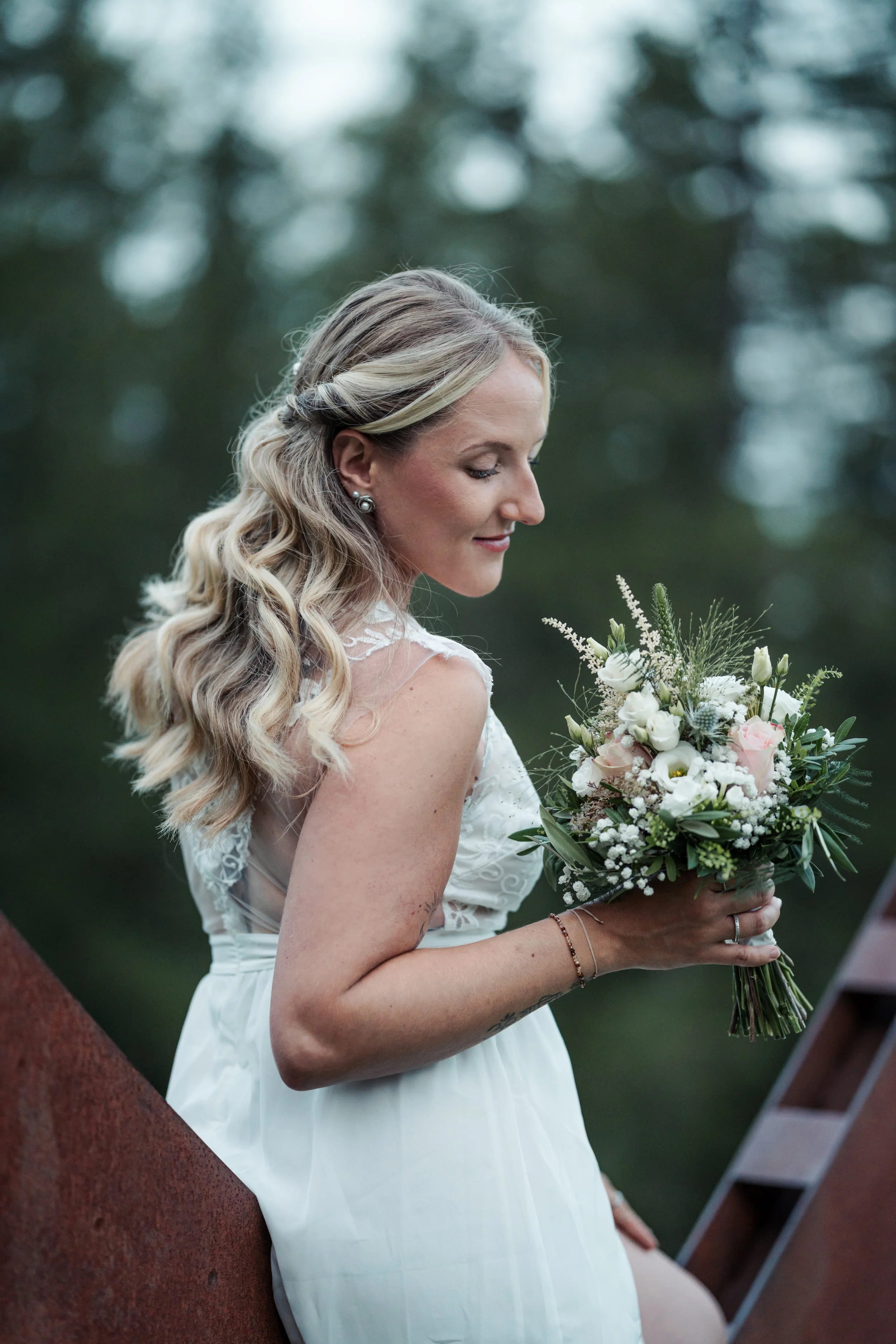 Junge Frau mit blonden, gewellten Haaren in Hochzeitskleid, hält Blumenstrauß, im Profil, im Freien, unscharfer grüner Hintergrund.