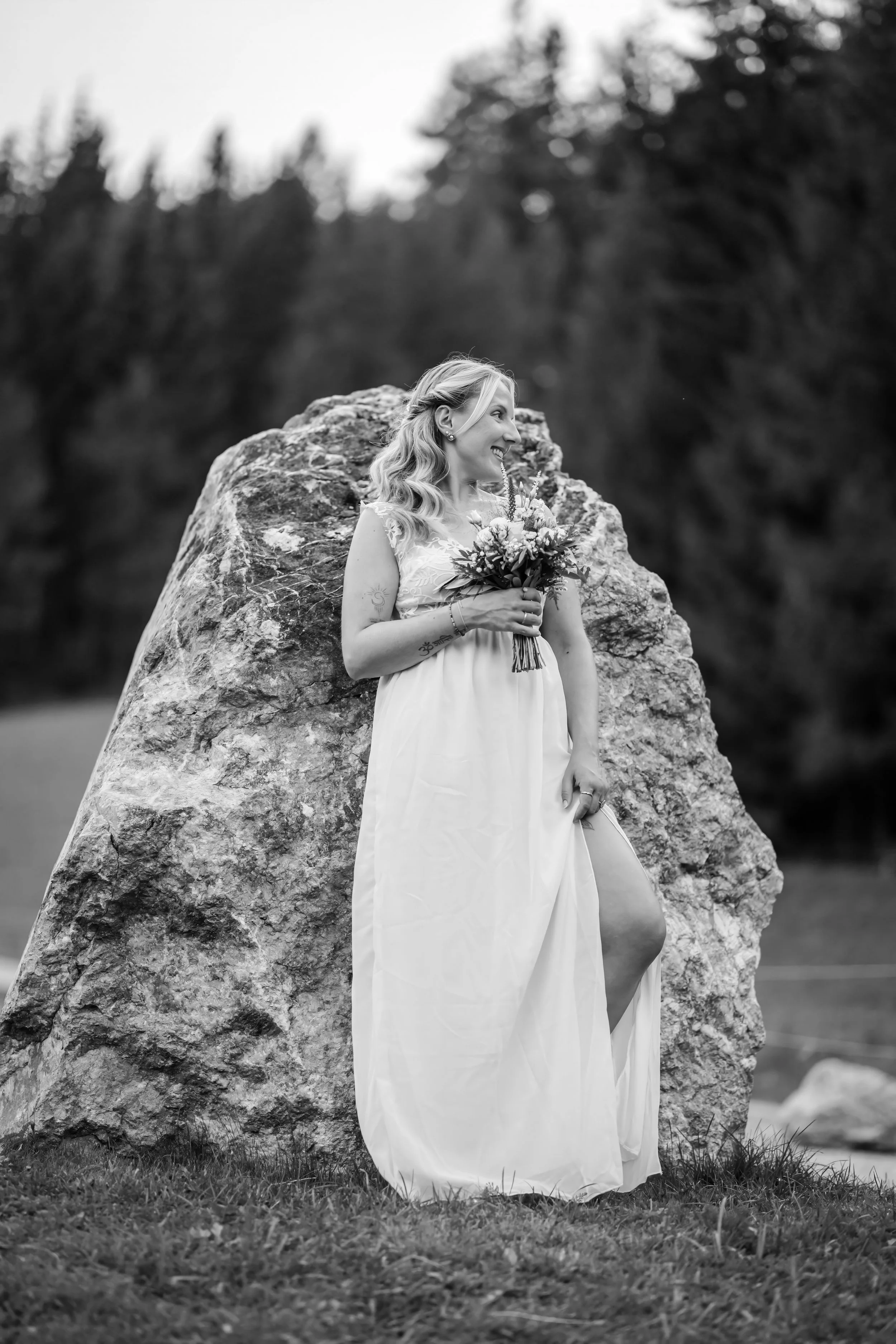 Eine Frau in einem weißen Kleid hält einen Blumenstrauß und steht vor einem großen Felsen im Freien, umgeben von Gras und Bäumen, im schwarz-weißen Bild.