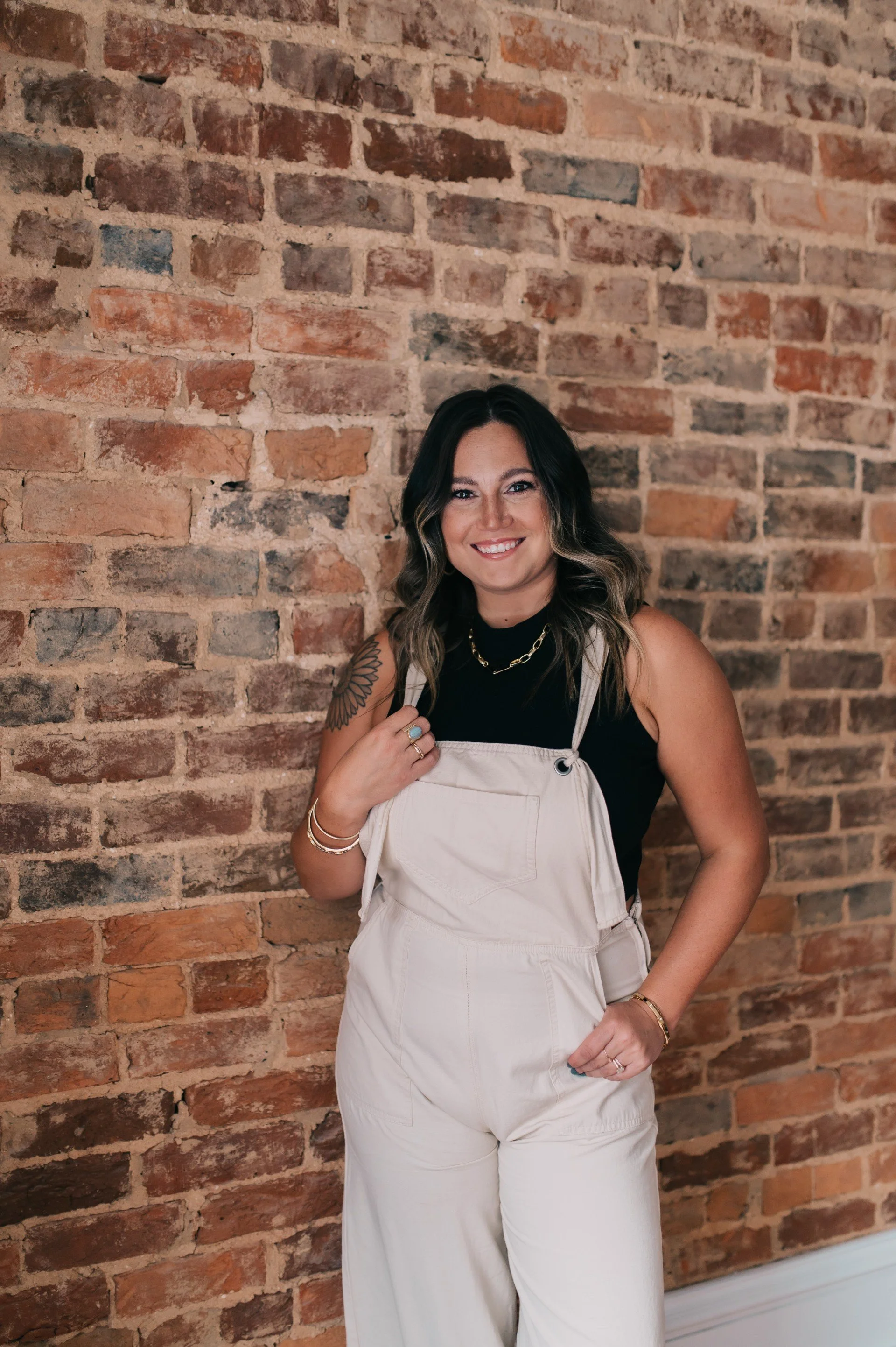 A woman with shoulder-length wavy dark hair and light skin, smiling and wearing a black sleeveless top, large gold chain necklace, and cream-colored overalls, standing against a red brick wall.