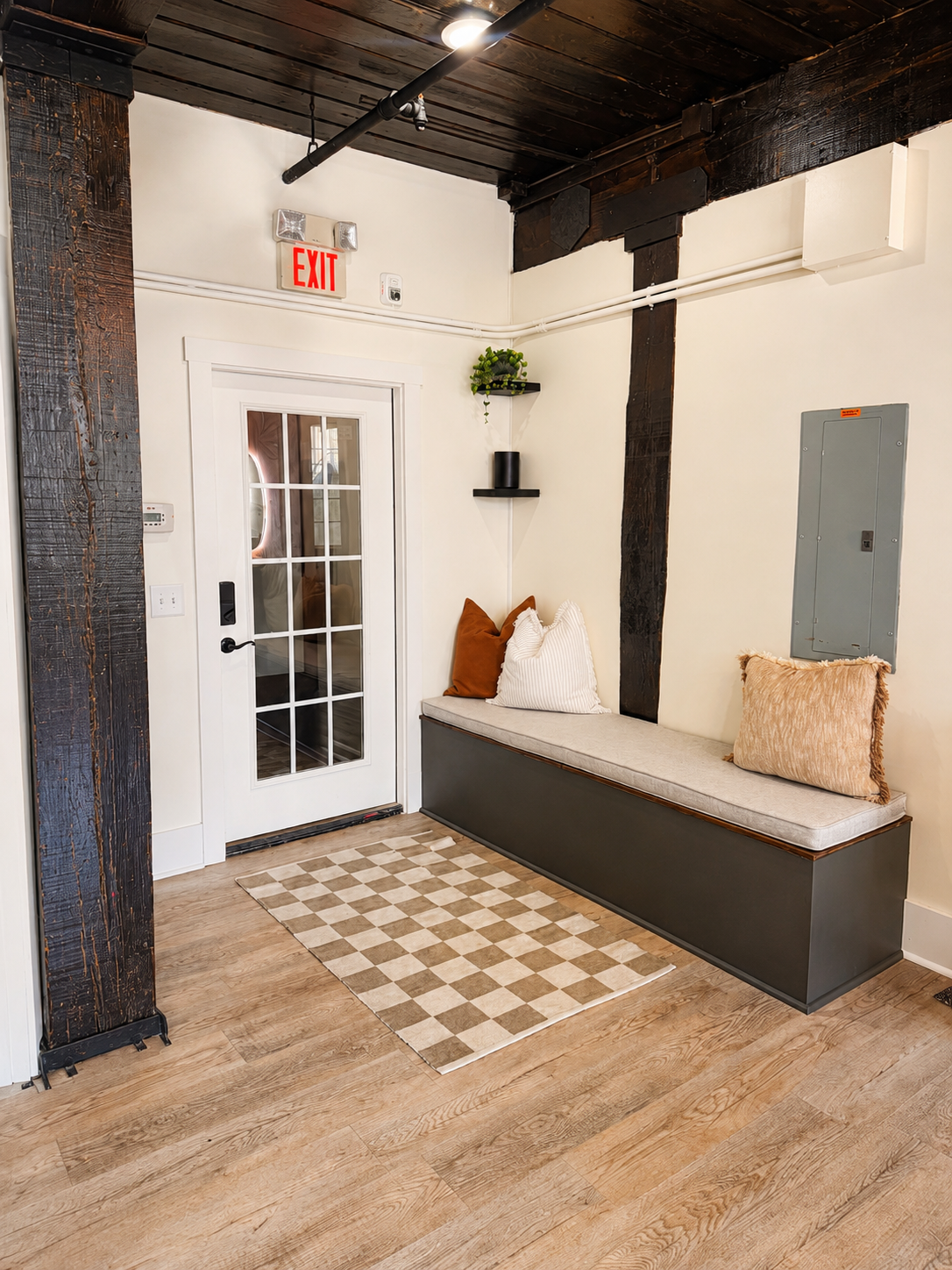 A cozy indoor nook with a cushioned seating area against the wall, decorated with pillows, next to a white door with glass panes, wooden flooring, a checkered rug, and black shelves with plants.