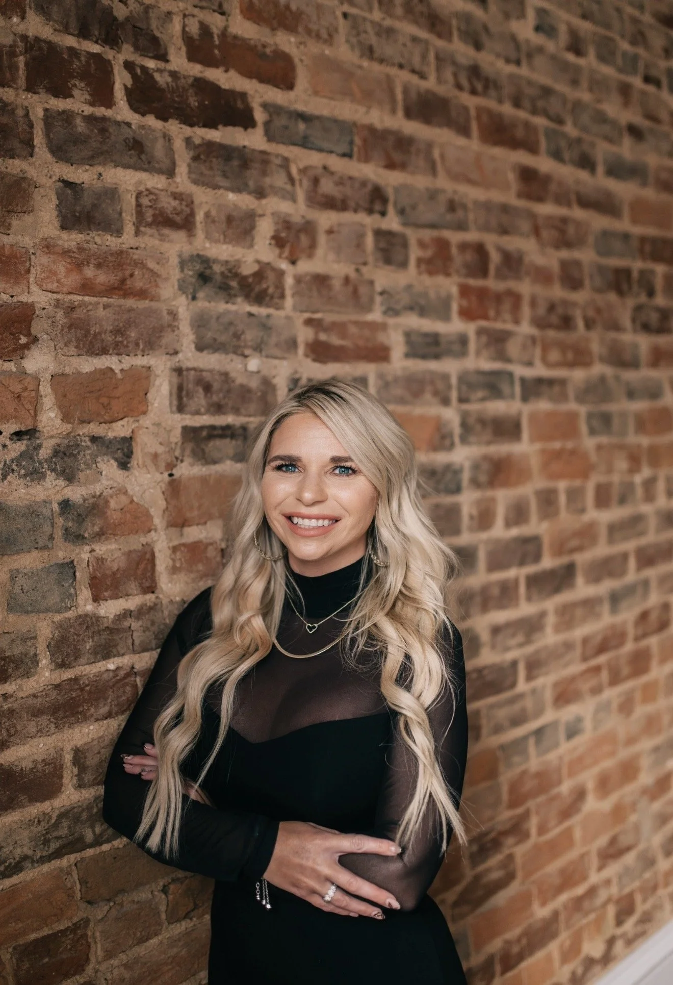 A woman with long blonde wavy hair, smiling, stands against a brick wall. She is wearing a black outfit with sheer details and layered necklaces.