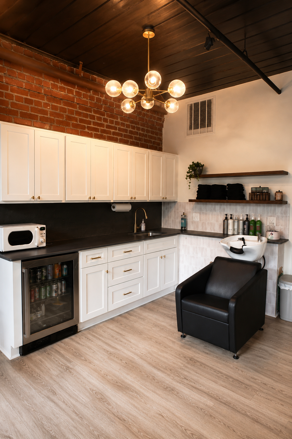 A modern salon or spa room with a salon sink, black chair, white cabinets, and a mini fridge, in a space with wooden flooring, a brick wall section, and modern lighting.