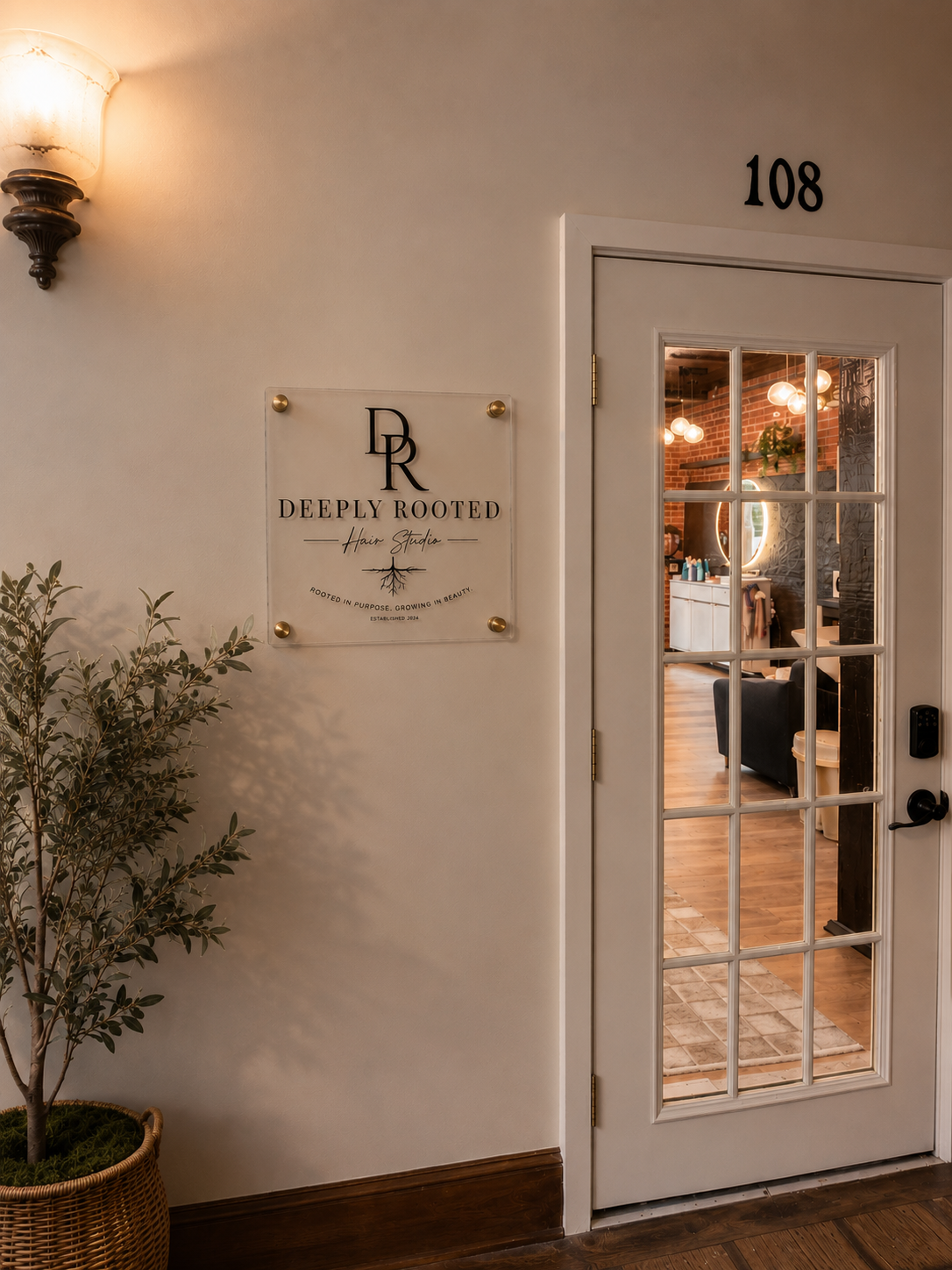 Interior view of a hair studio with a door labeled '108', a sign reading 'Deeply Rooted Hair Studio', and a potted plant next to it, showing a cozy salon with furniture and lighting inside.