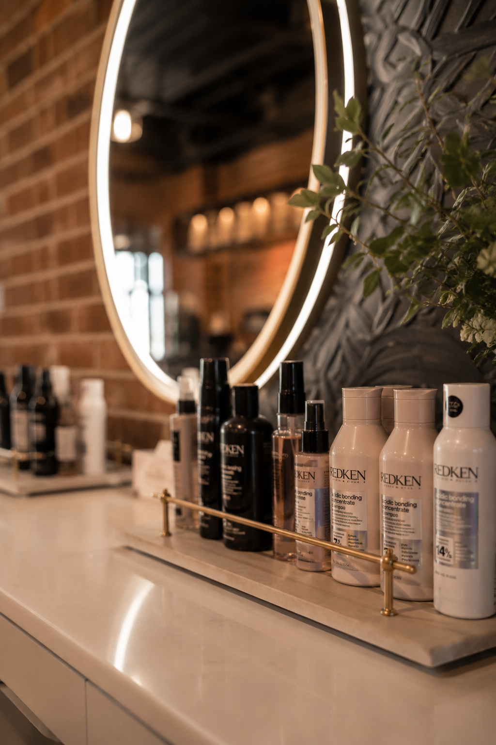 Beauty salon countertop with bottles of Redken shampoo and styling products, large illuminated mirror, and greenery in the background.