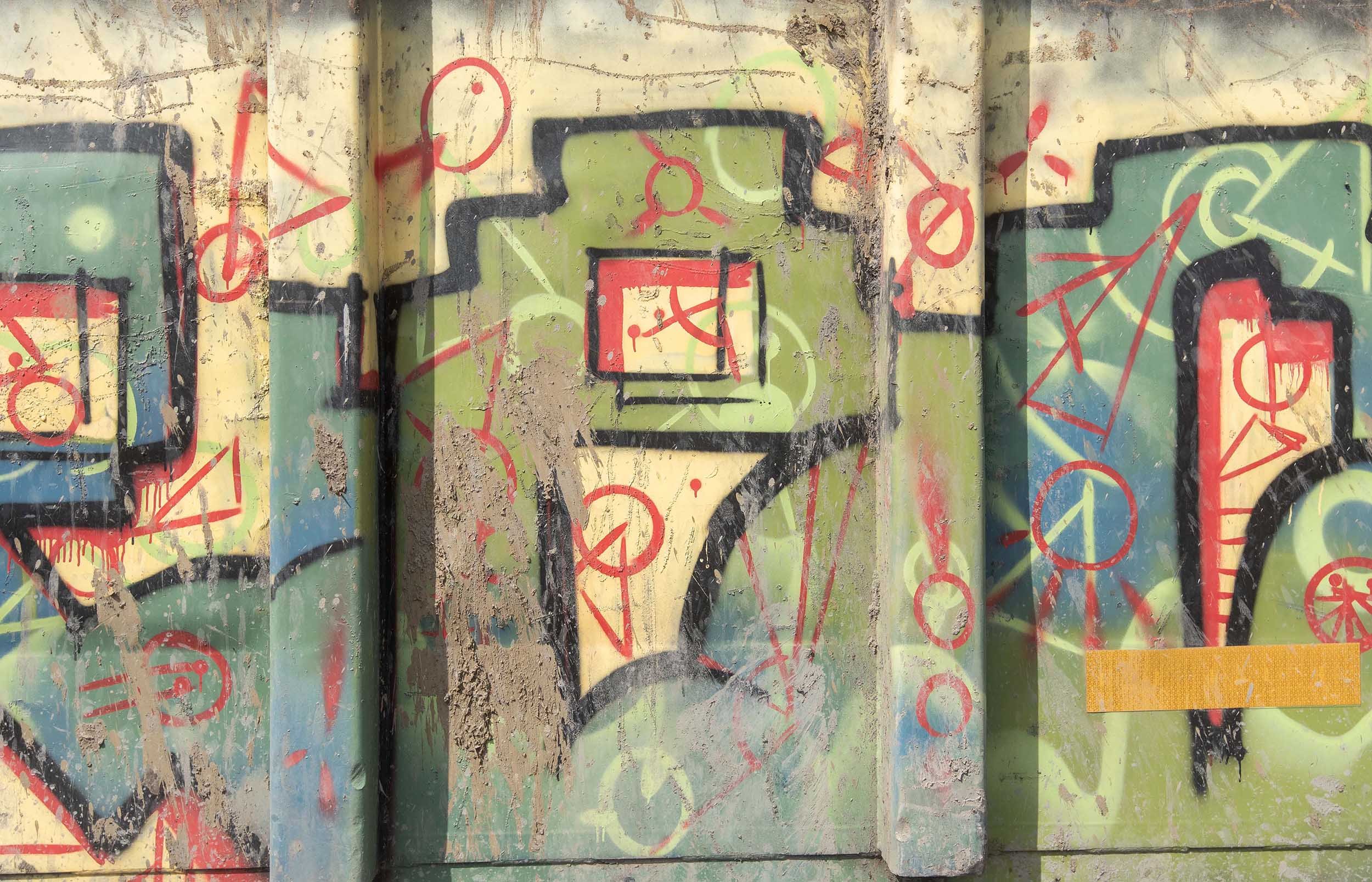 Close-up of a section of graffiti art on a weathered wall with multiple colors including red, black, green, yellow, and blue. The graffiti features abstract shapes, lines, and numbers over a textured surface with chipped paint.