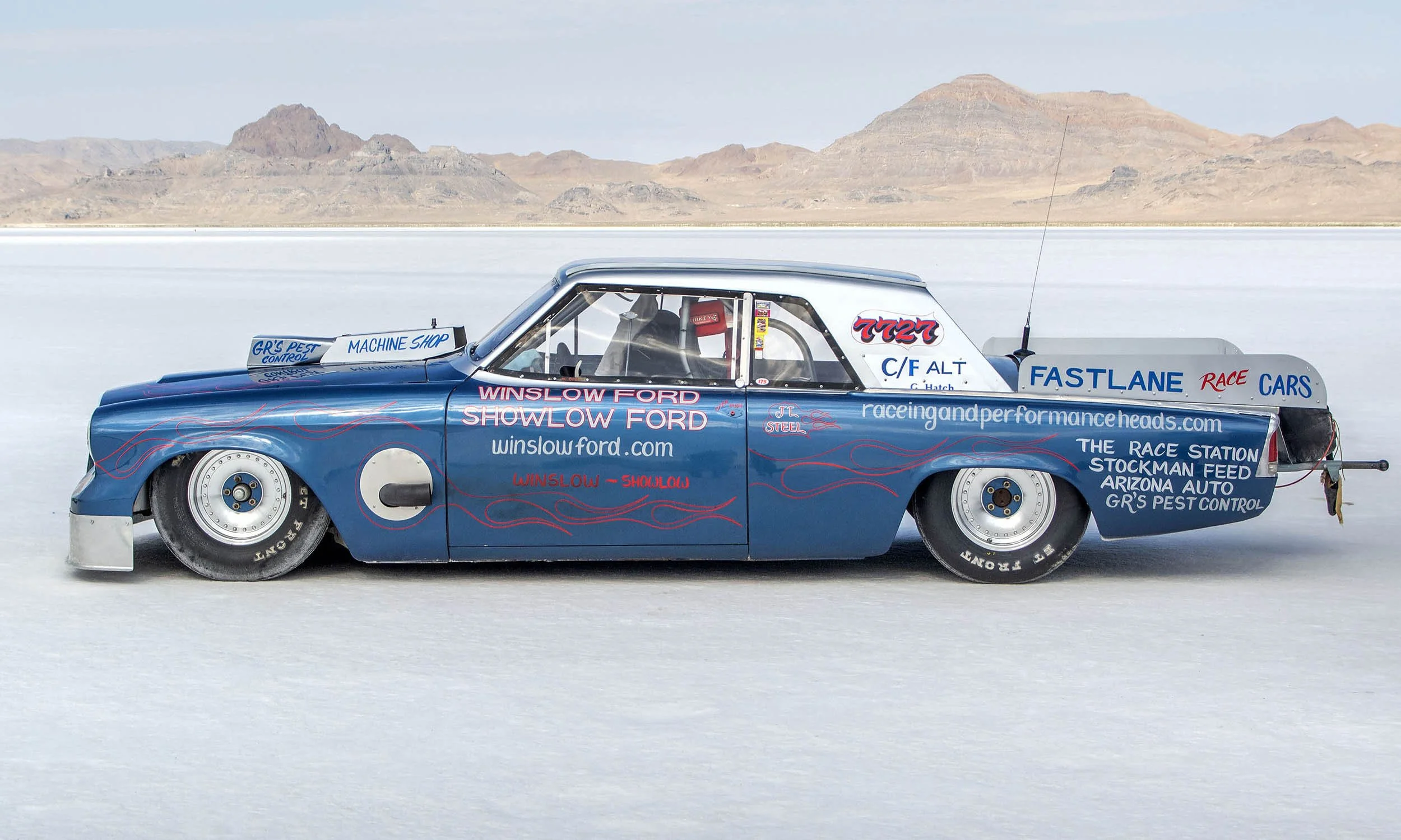 A vintage blue race car with red flame decals is parked on a salt flat with mountains in the background. The car is covered with various sponsor decals and website URLs, including 'winslowford.com'.