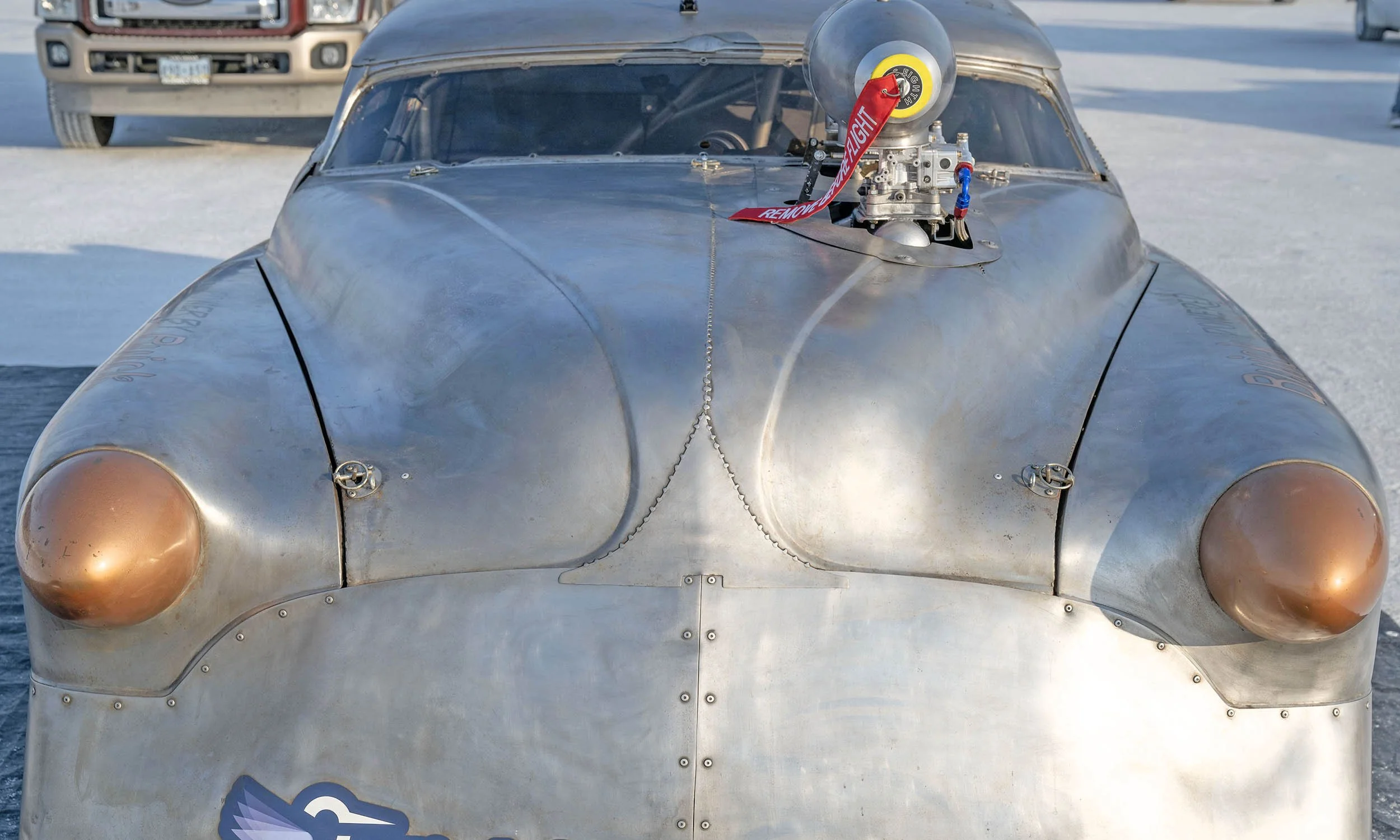 A vintage silver race car with a large supercharger on the hood and gold-colored headlights. A red strap with the words 'REMOVE BEFORE FLIGHT' is attached to the supercharger. The car is on a snowy surface, with another vehicle visible in the background.