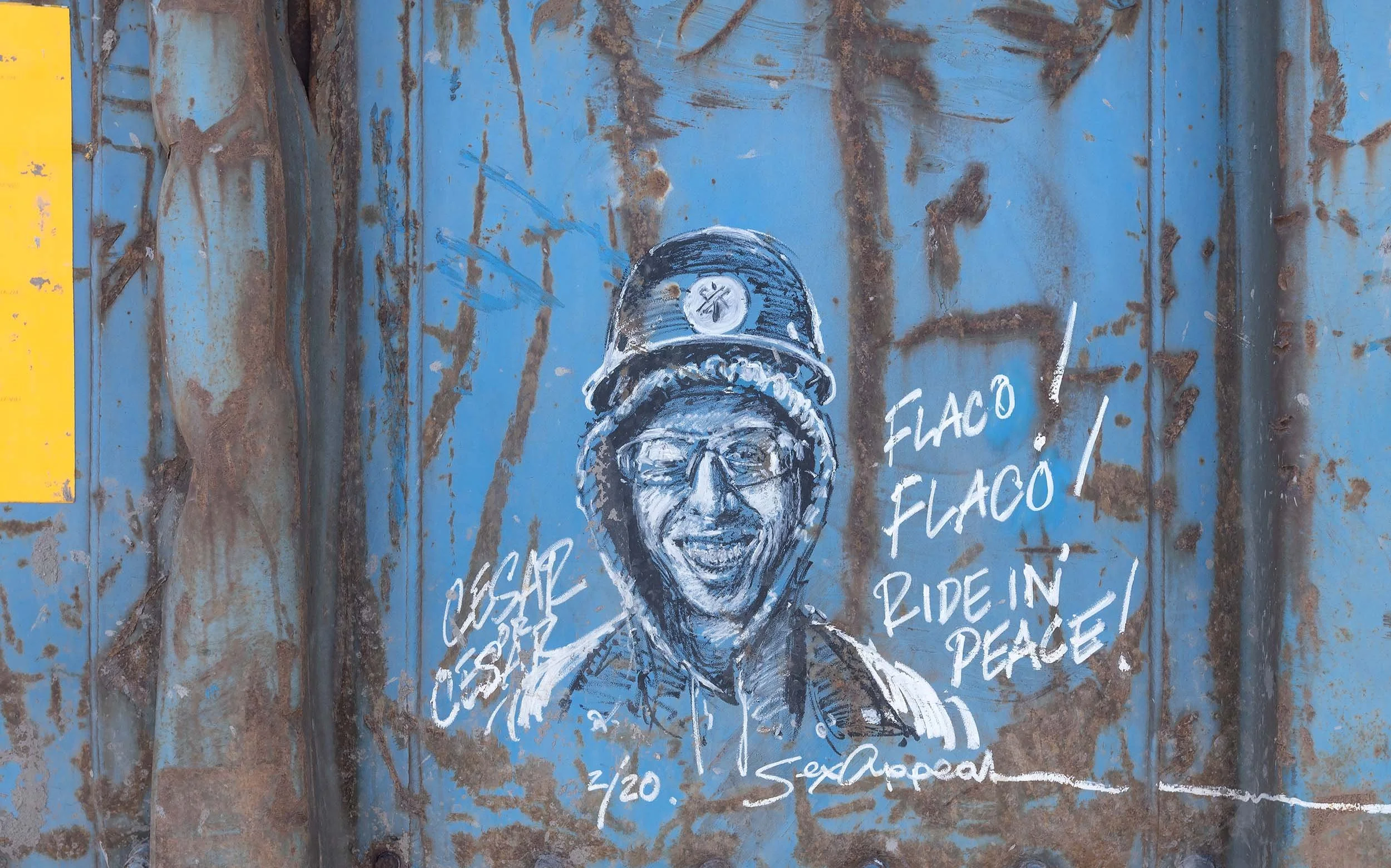 A graffiti mural featuring a smiling man wearing a helmet and glasses, with handwritten phrases such as 'FLACO,' 'RIDE IN PEACE,' and others on a rusty, blue metal surface.