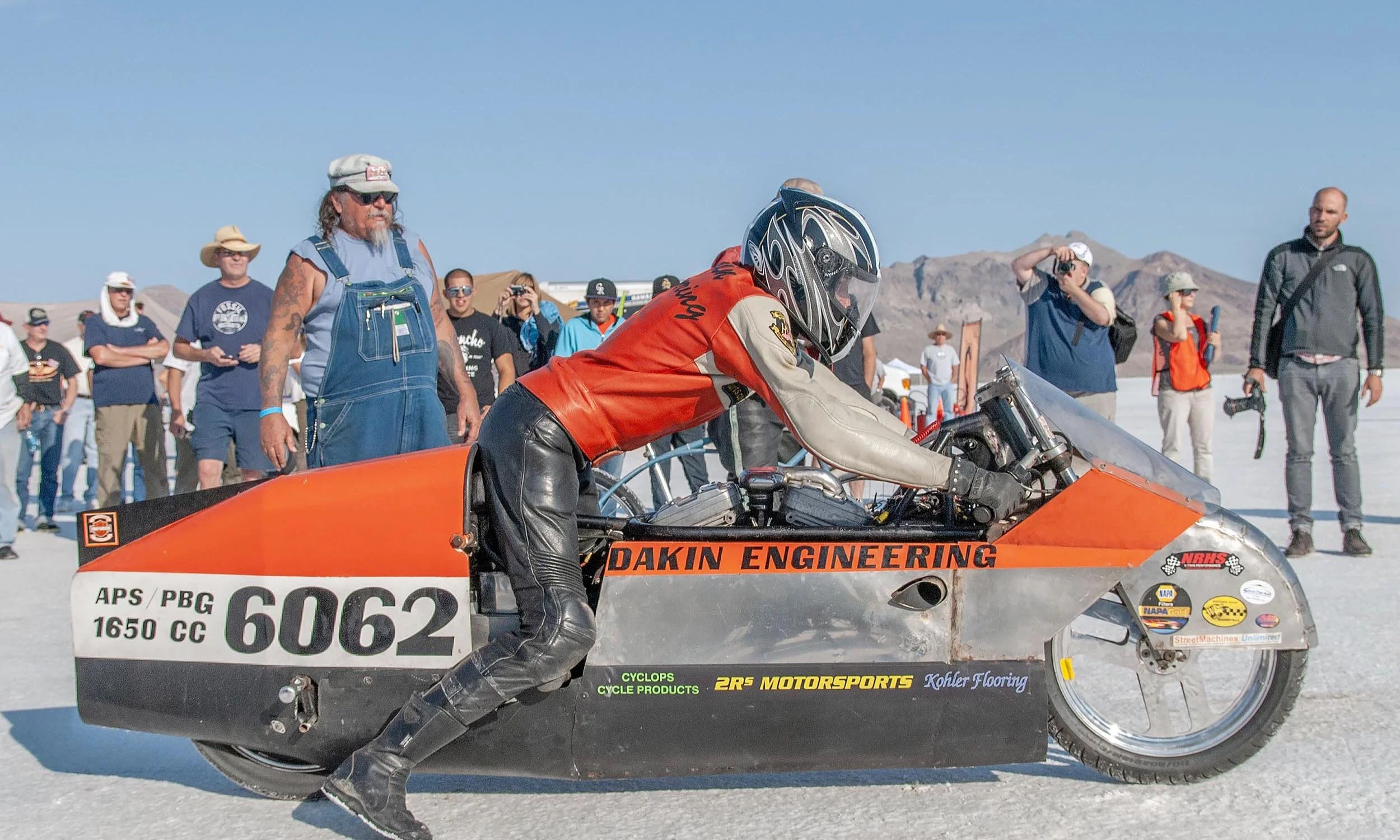 A person in racing gear and a helmet preparing a drag racing vehicle with sponsors and labels, while a group of spectators and photographers watch in the background in a desert setting with mountains.