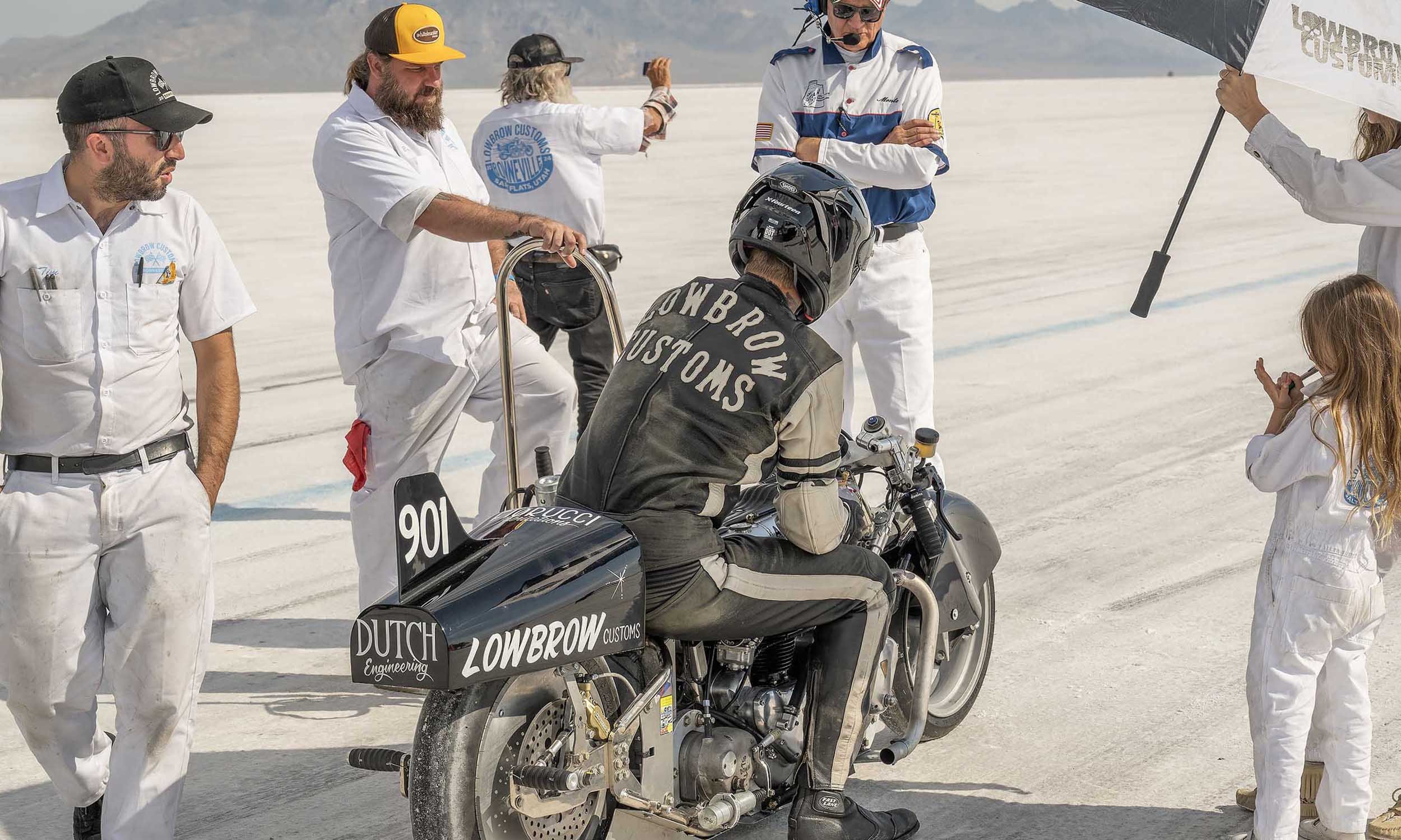 A group of people in white jumpsuits and a person in motorcycle gear gather around a black motorcycle on a salt flat. The motorcycle has various sponsor stickers, including Dutch Lowbrow Customs. Some individuals wear helmets, and others hold umbrell