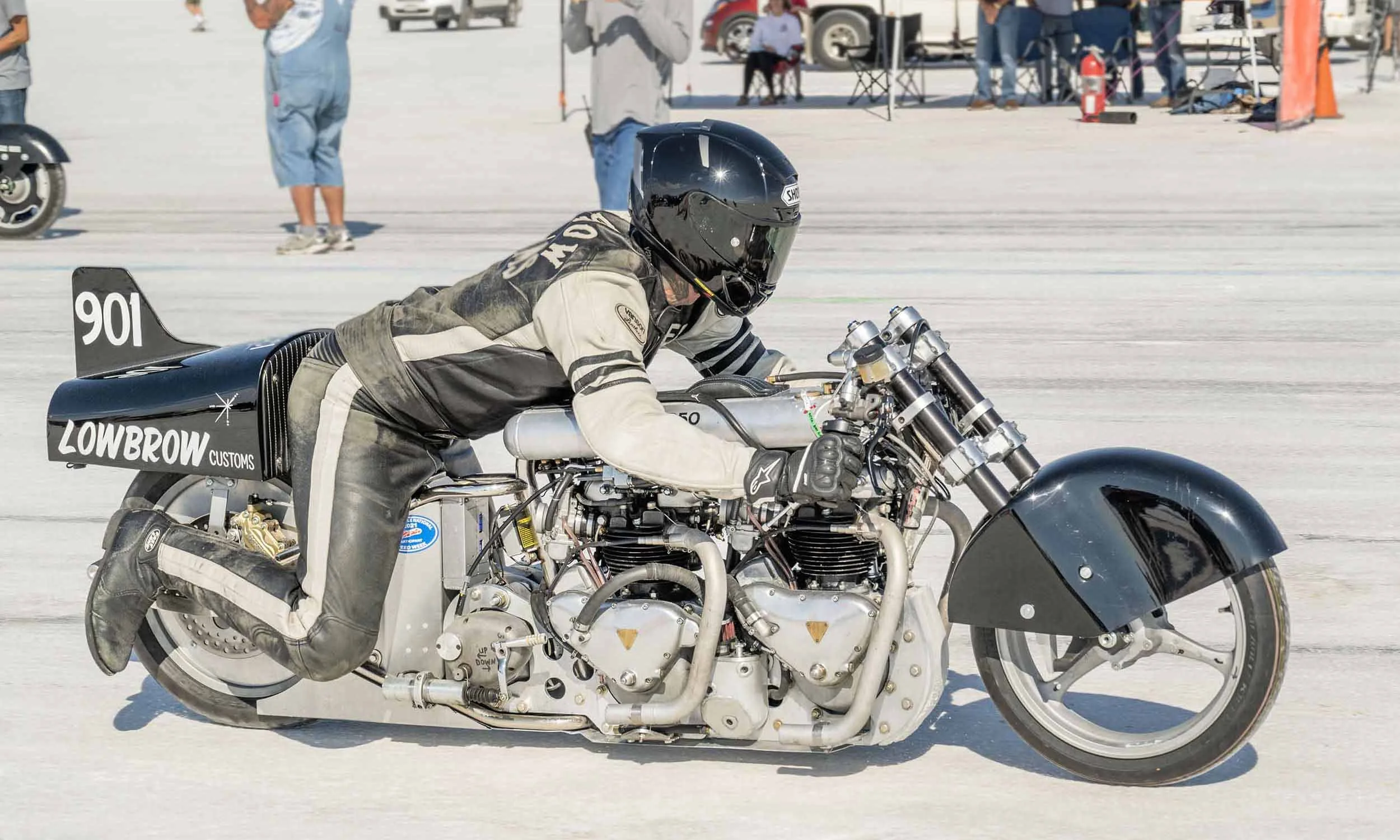 A person in a racing suit and helmet riding a custom lowbrow-style motorcycle on a flat, salt surface at a racing event, with spectators and vehicles in the background.