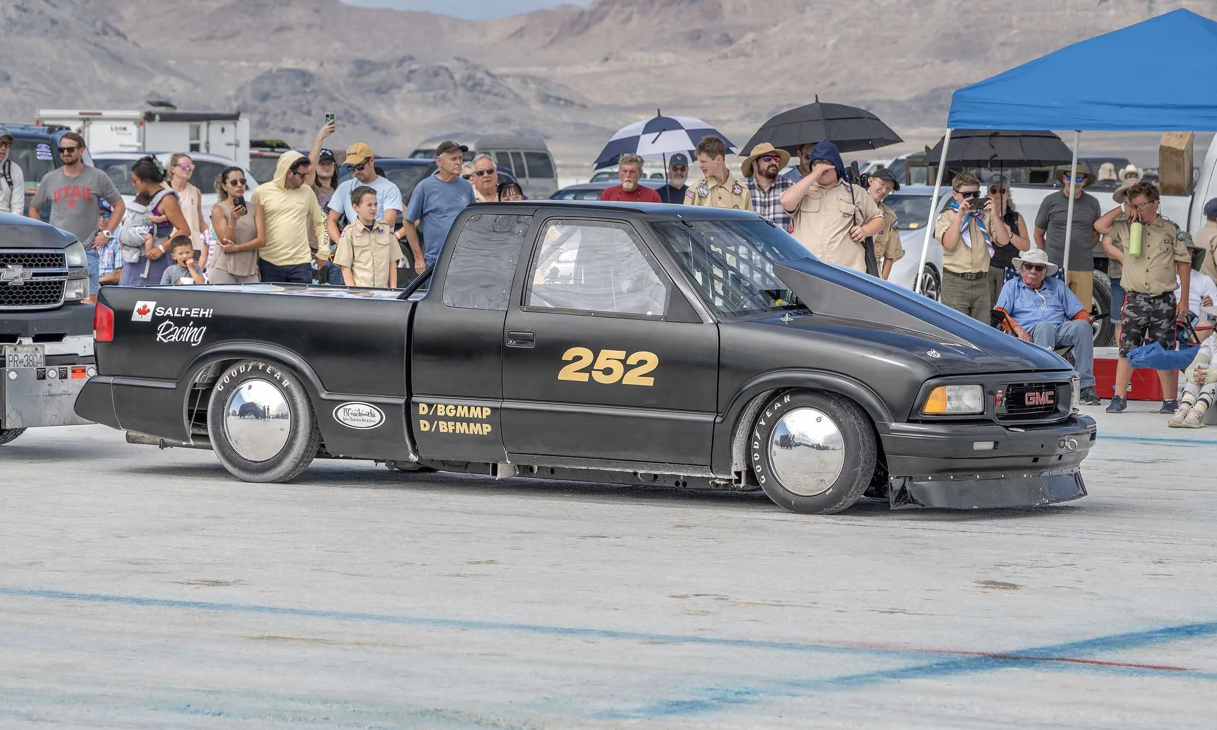 A black race car with the number 252 and various sponsor decals, parked on a race track with a crowd of spectators and people in scout uniforms standing behind it, with mountains in the background.