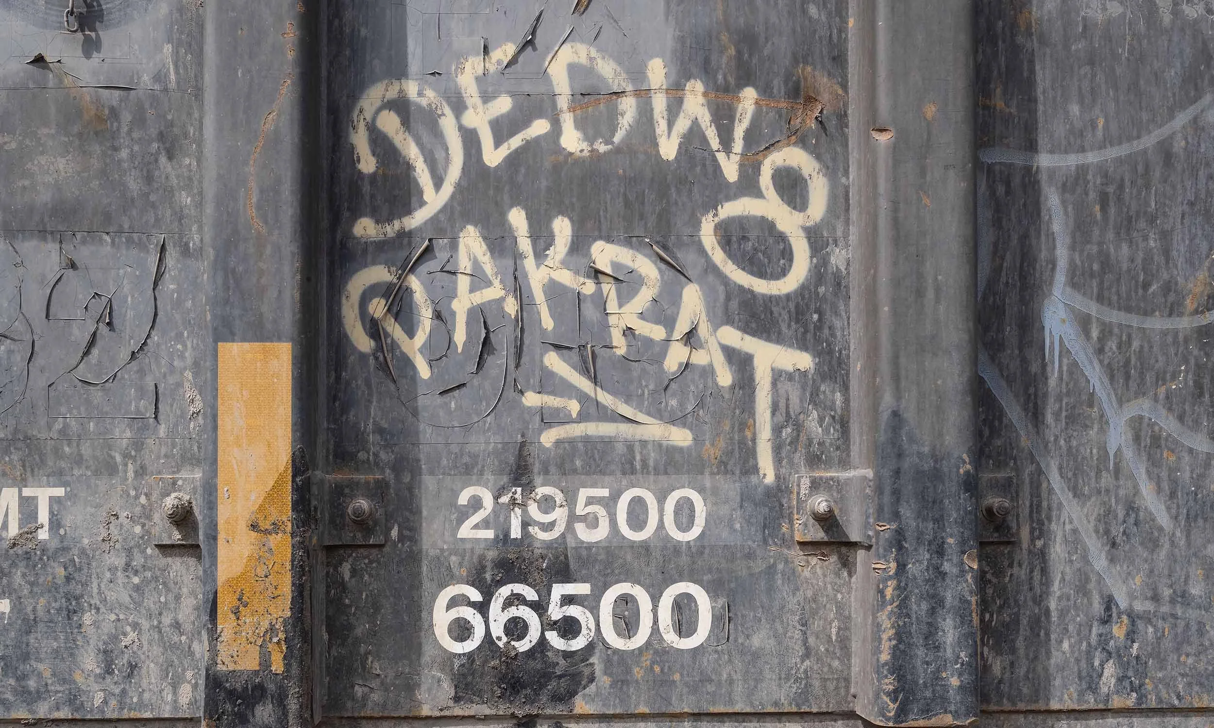 Close-up of a weathered metal surface with peeling paint and graffiti. The graffiti reads 'DUSTW 0' in large, white spray paint. There are rust marks and remnants of other painted markings visible.