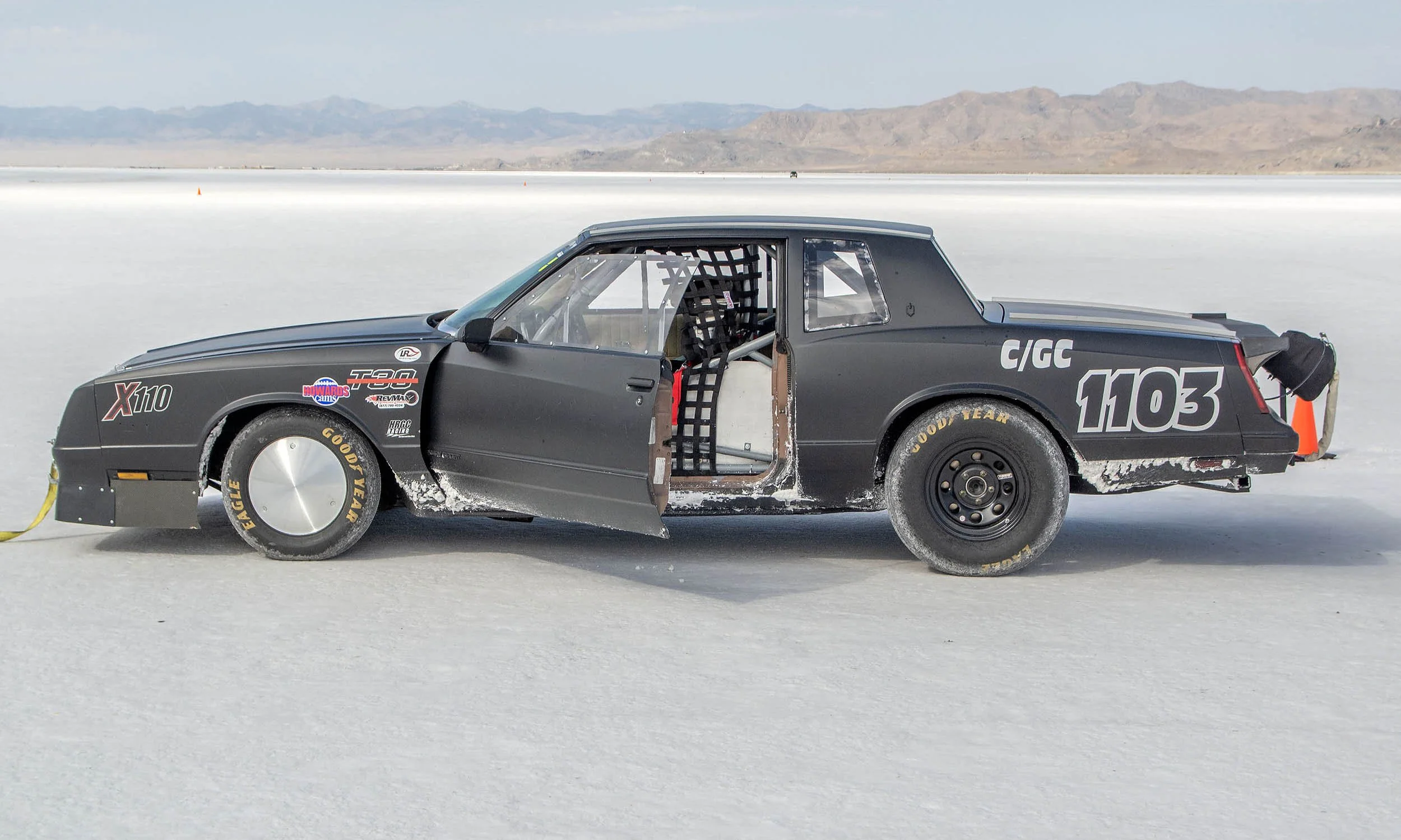 A black race car with racing decals and the number 1103 on the side, sitting on a salt flat with mountains in the background.