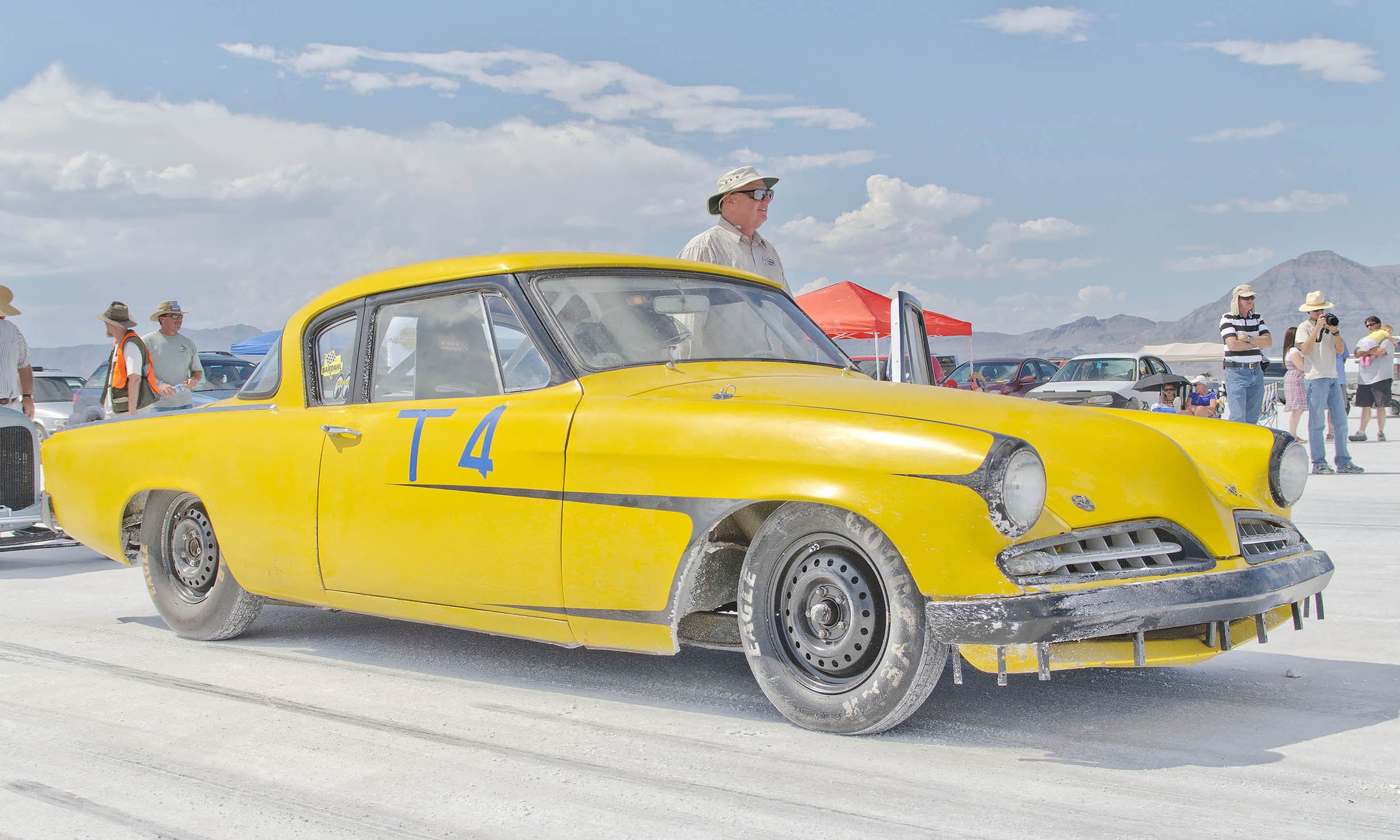 Vintage yellow race car with the number T4 on the side at a racing event, surrounded by people in hats and sunglasses, some taking photos.