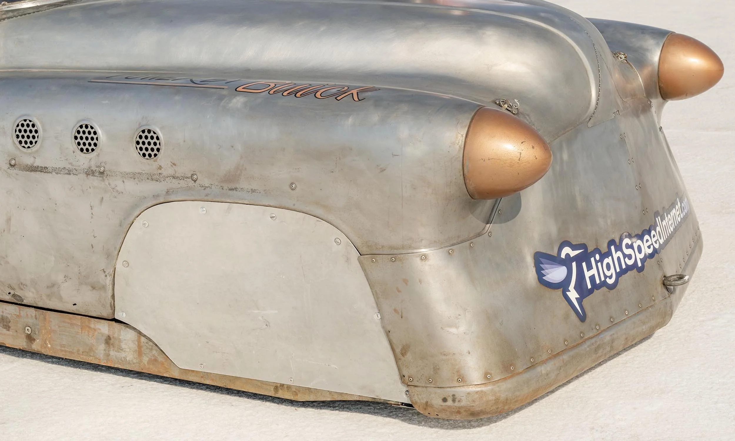 Close-up of a vintage silver race car with copper accents, showing the front wheel arch, grille vents, and a sticker saying 'HighSpeedLink.com' on the side.