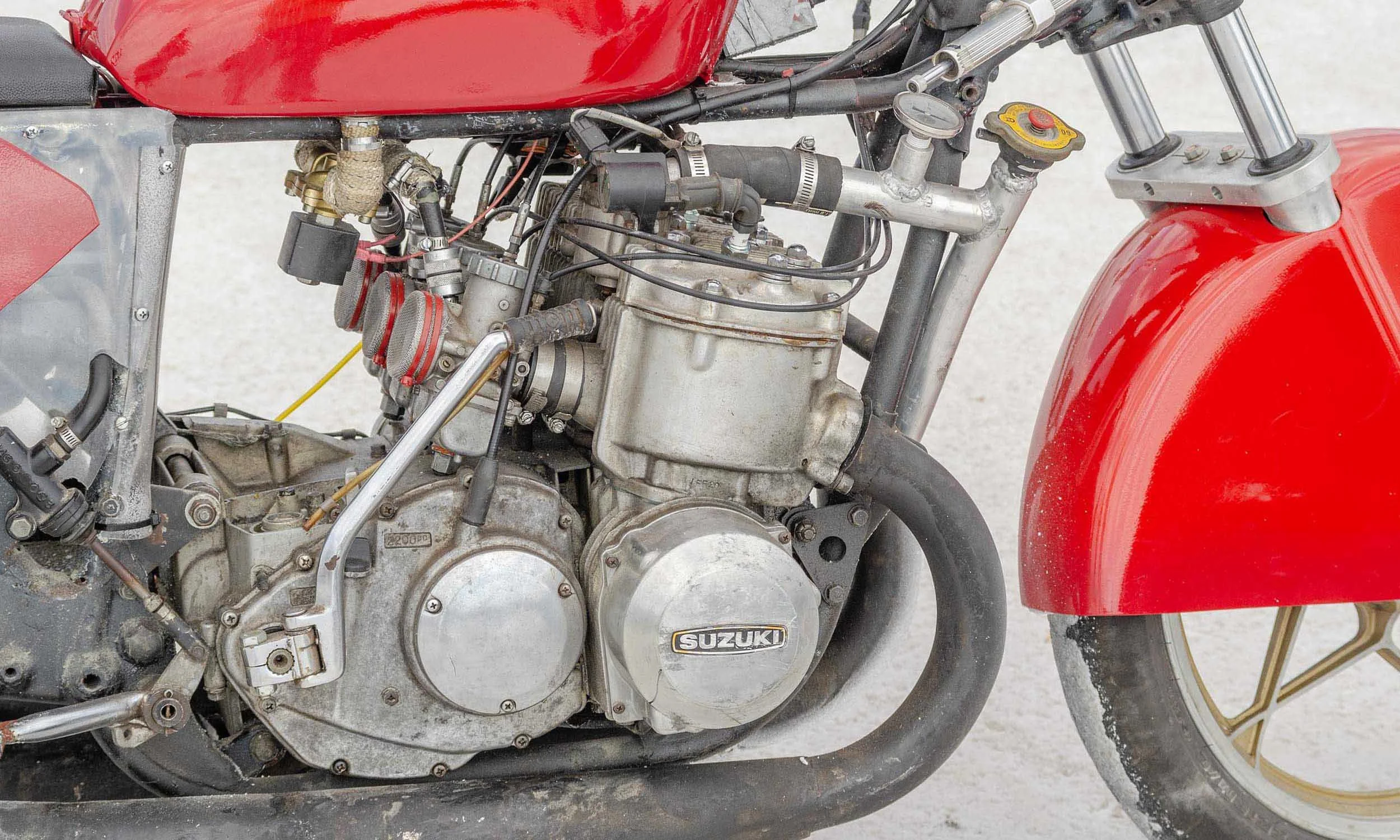 Close-up of a motorcycle engine with a red fuel tank and partial red front fender, showing the Suzuki logo on the engine cover, with visible wires, hoses, and mechanical components.