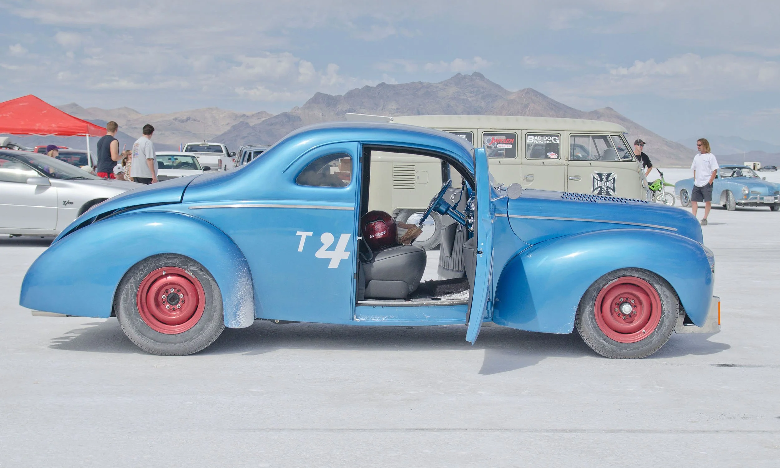 A vintage blue race car with red wheels and the number 'T4' on its side in a parking lot, with people walking around and mountains in the background under a partly cloudy sky.