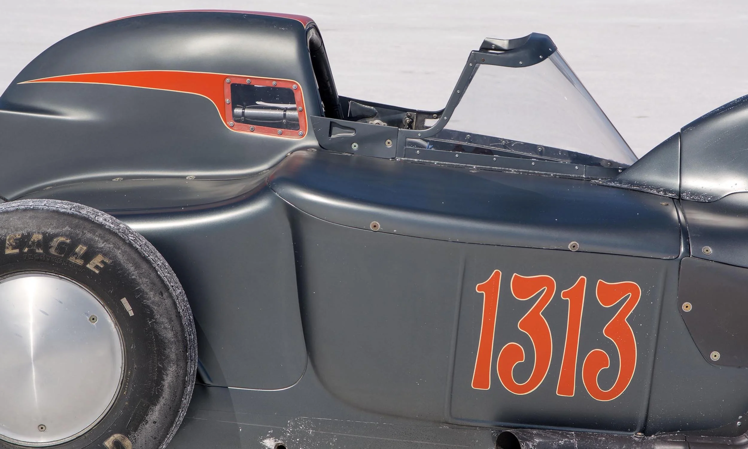 Close-up of a vintage race car showing the side window, part of the hood, front tire, and orange racing numbers on a gray body. The car is on a flat, icy surface.