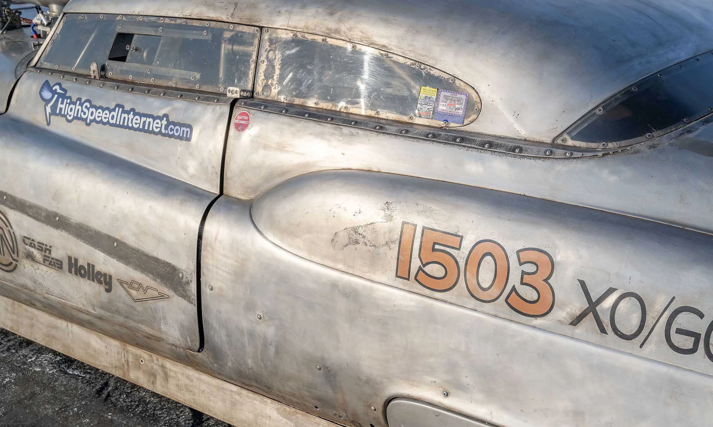 Close-up of a metal race car with various stickers and decals, including a HighSpeedInternet.com logo, a sticker indicating an electric disconnect, and several racing regulation stickers. The car's surface is weathered and metallic, with rivets and p