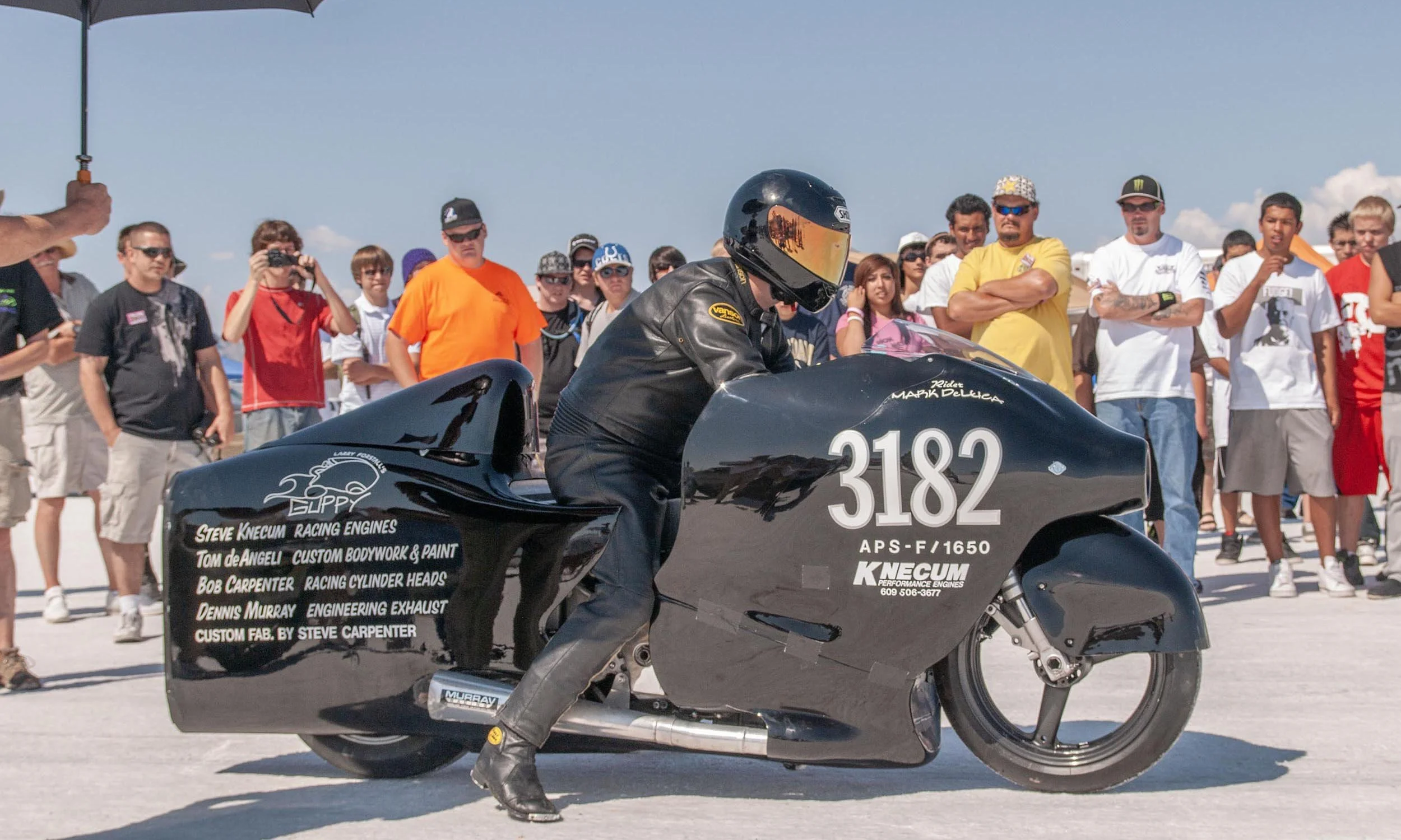 A person wearing a black helmet and leather jacket is sitting on a black racing motorcycle at a racing event. Behind him, a group of spectators, including men, women, and children, are watching. The motorcycle has various sponsor names and numbers on