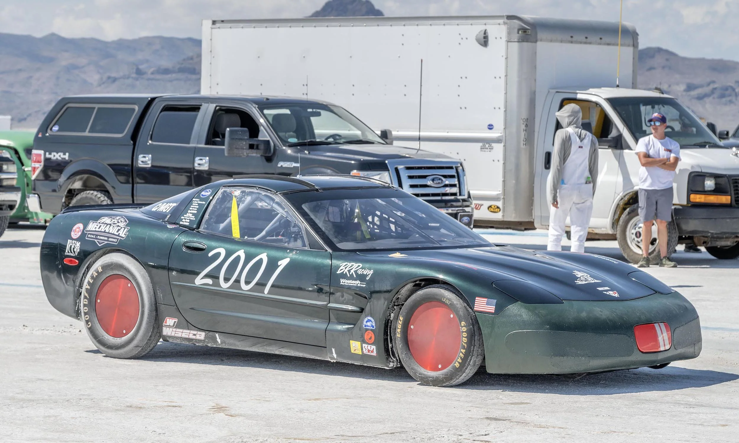 A black race car with the number 2001 on the side is on a race track, with a gray pickup truck and a white cargo van parked in the background. Two people are standing near the white van, one wearing white and the other wearing a gray hoodie and sungl